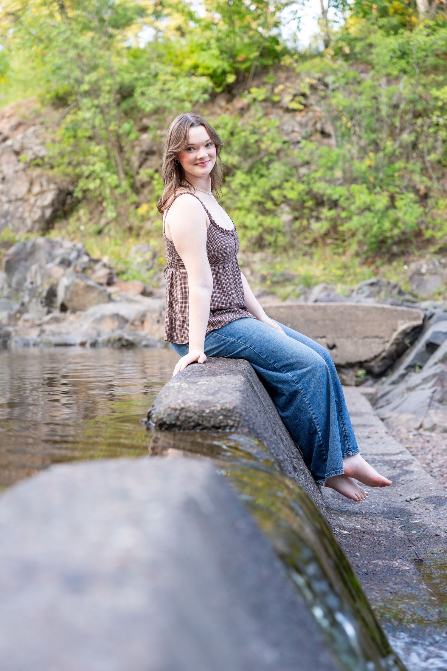 Young woman in jeans and brown top sits barefoot on rocks by a peaceful stream surrounded by lush greenery.