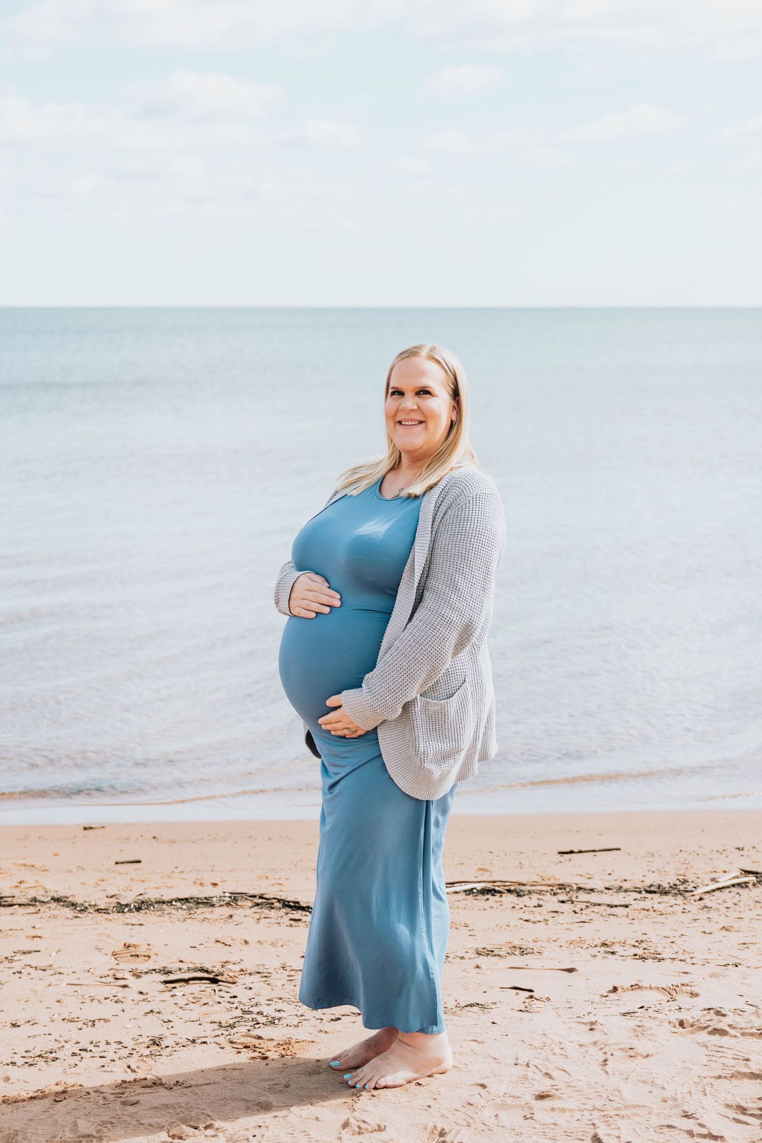An expectant mother in a blue dress and gray cardigan stands on a beach touching her baby bump.