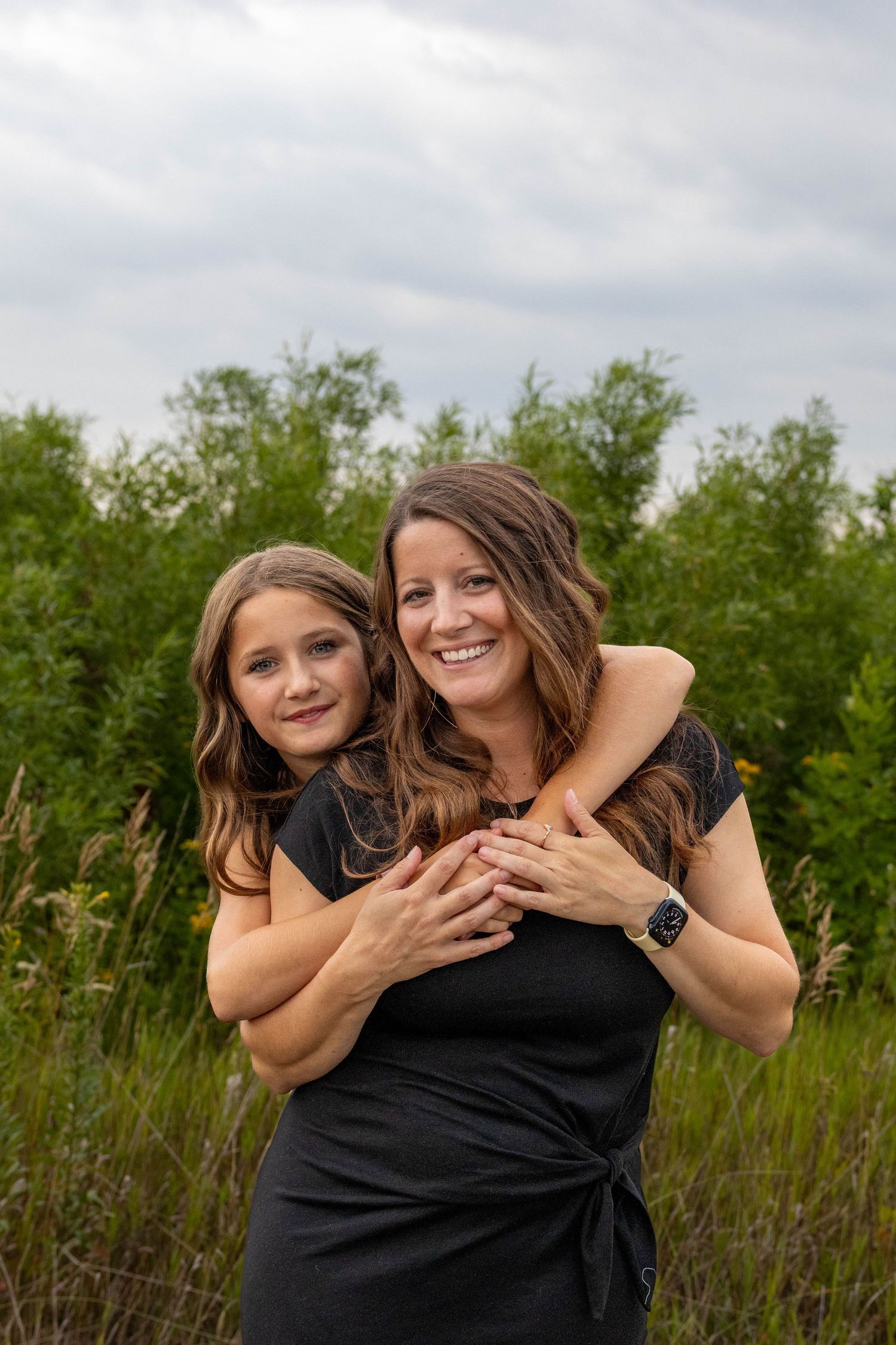 Two women in black dresses share joyful embraces while posing outdoors against a green natural background.