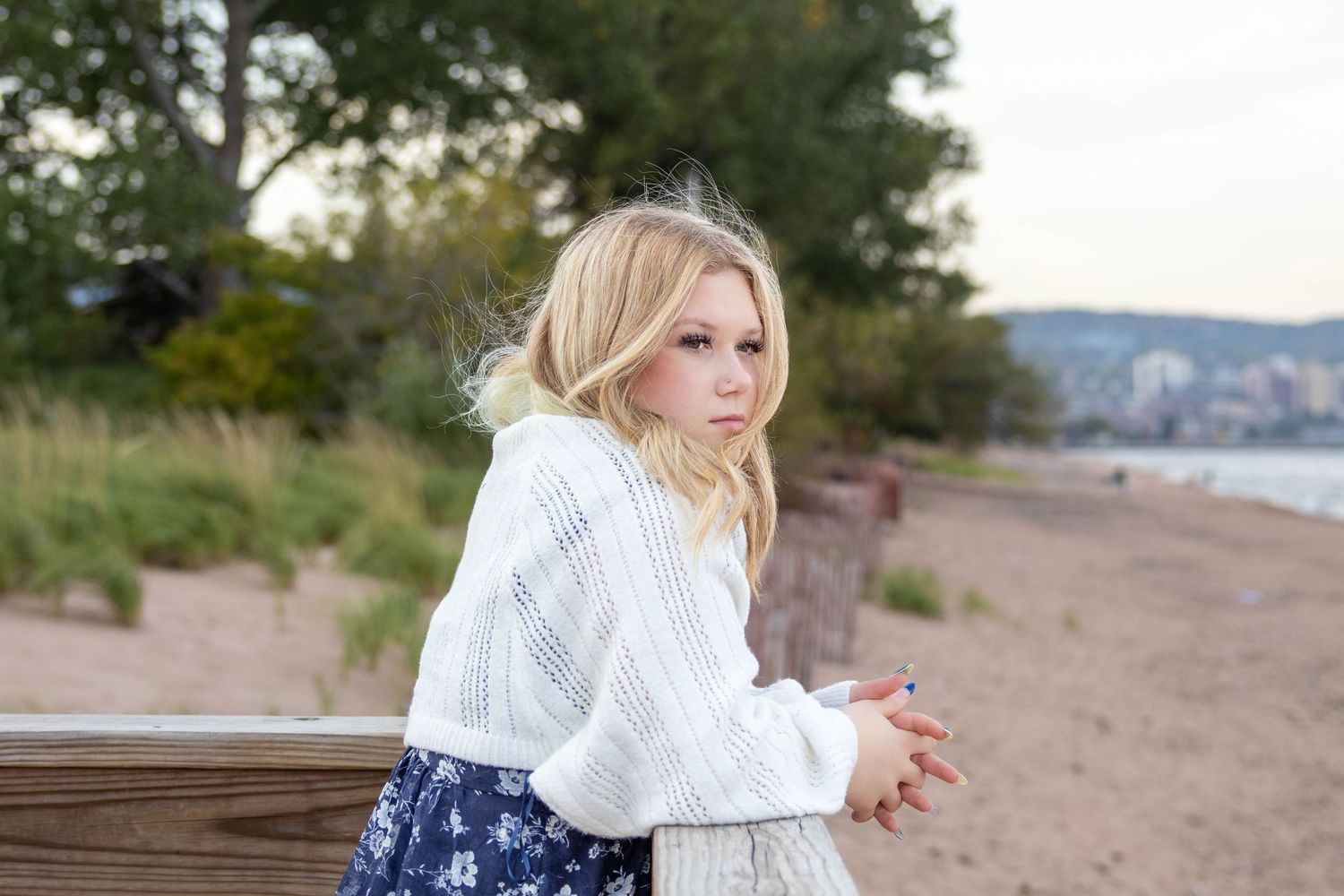 Person in white sweater sits thoughtfully on wooden bench at sandy beach during autumn day.