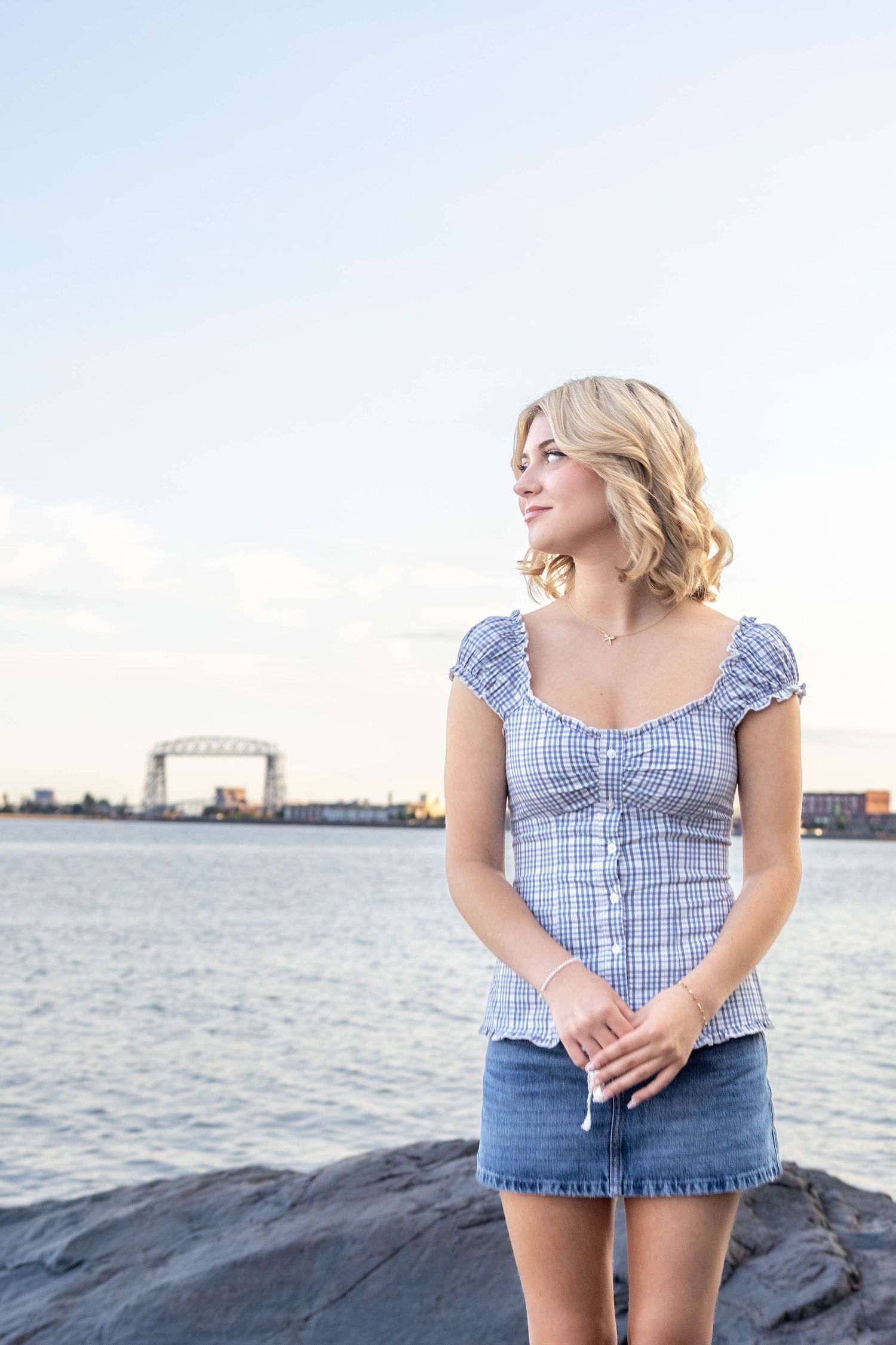 Woman in blue gingham top and denim shorts poses by waterfront during sunset.