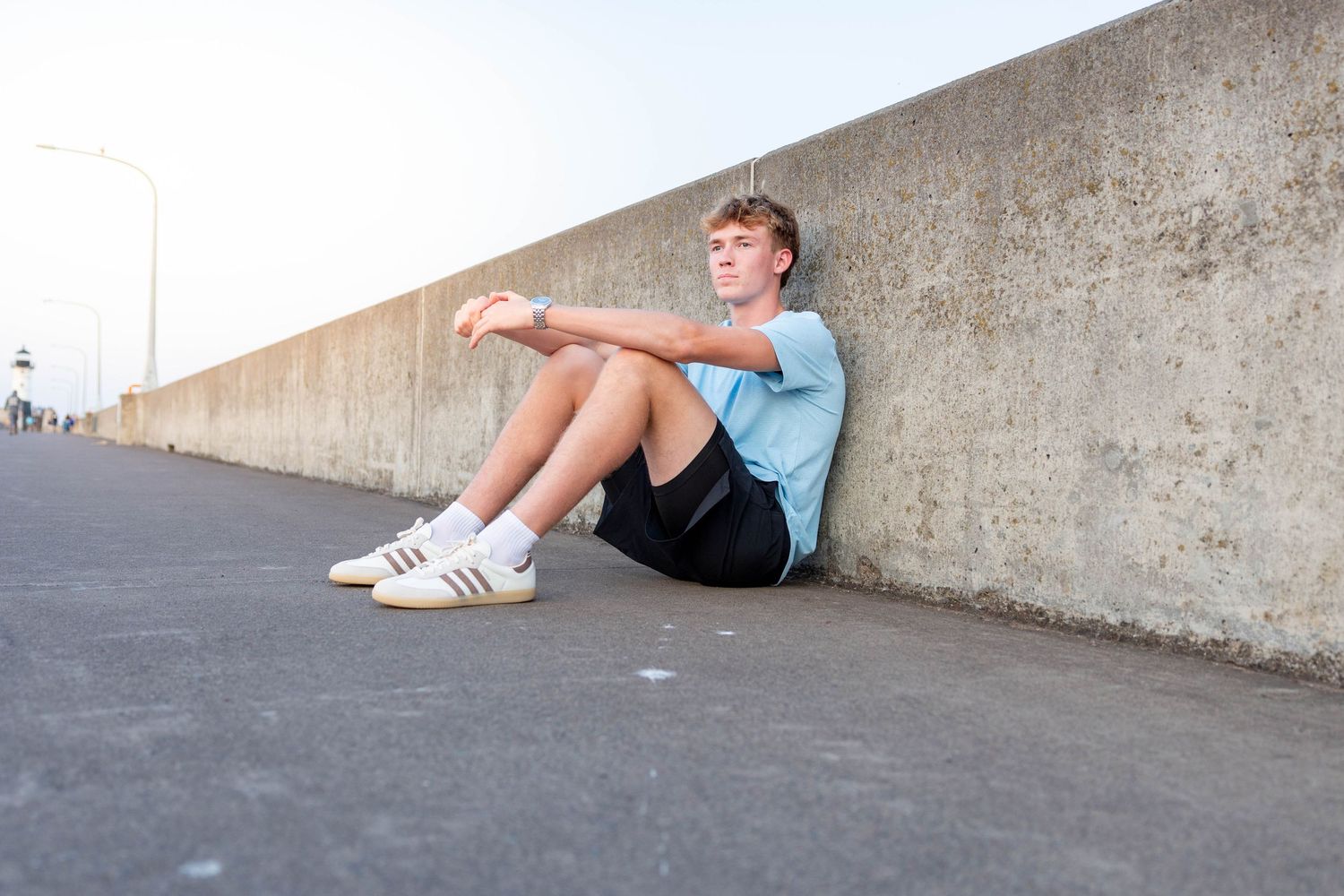 Teen boy sitting against concrete wall outdoors, wearing blue t-shirt, black shorts, and white Adidas sneakers.