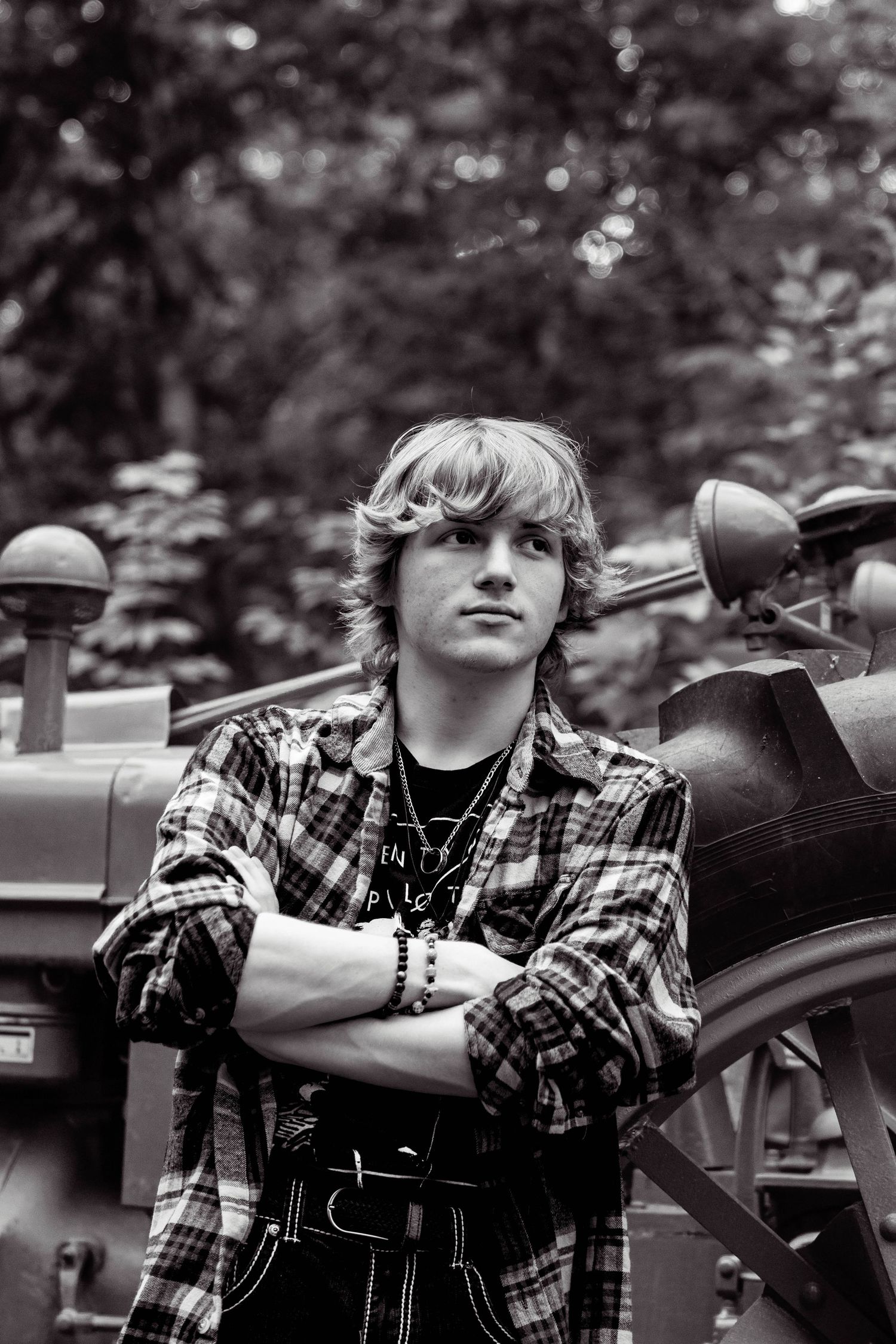 Black and white portrait of a person in a plaid flannel shirt leaning against a vintage motorcycle outdoors.