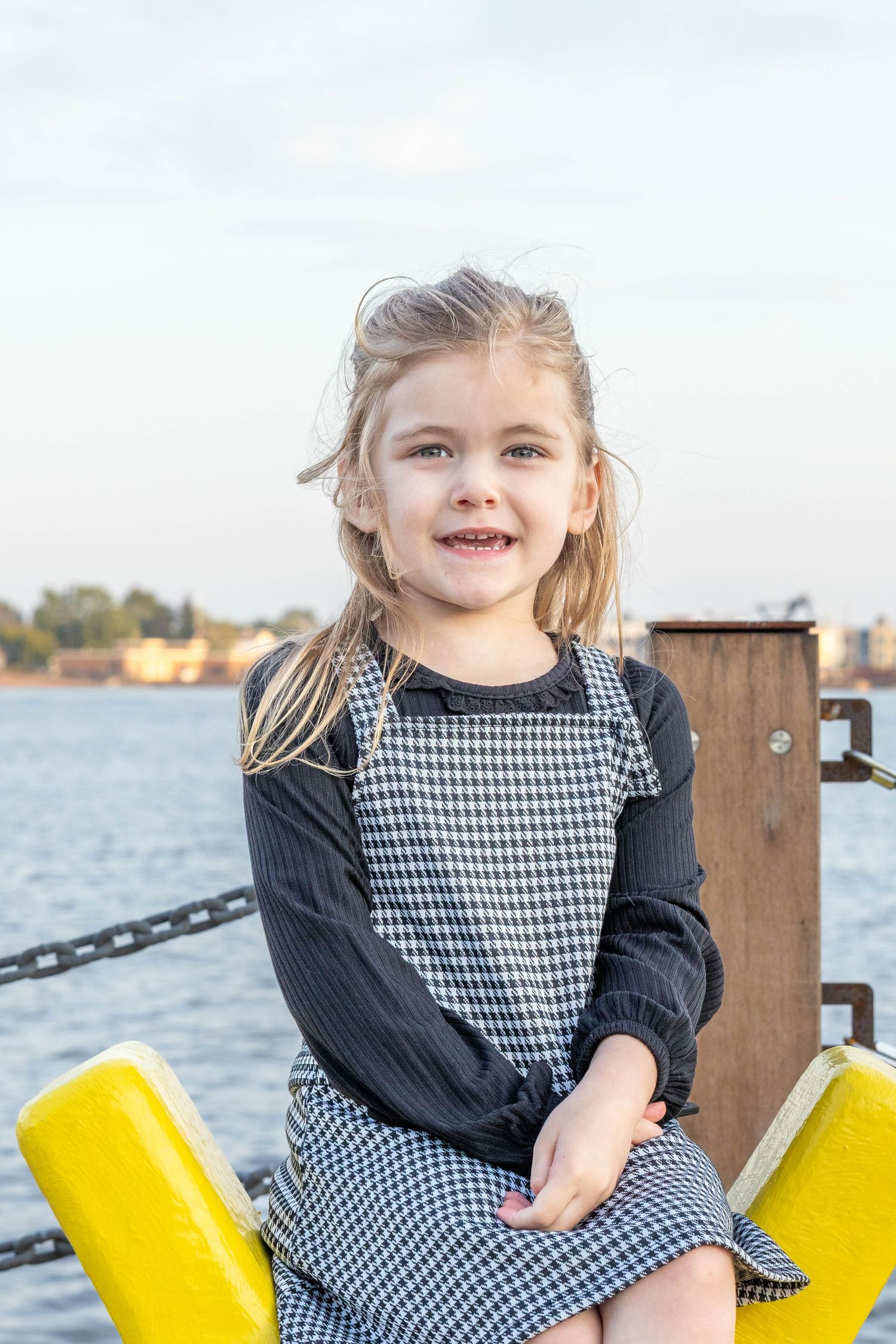 A child in a houndstooth dress and black sweater sits on a yellow bollard by the waterfront.