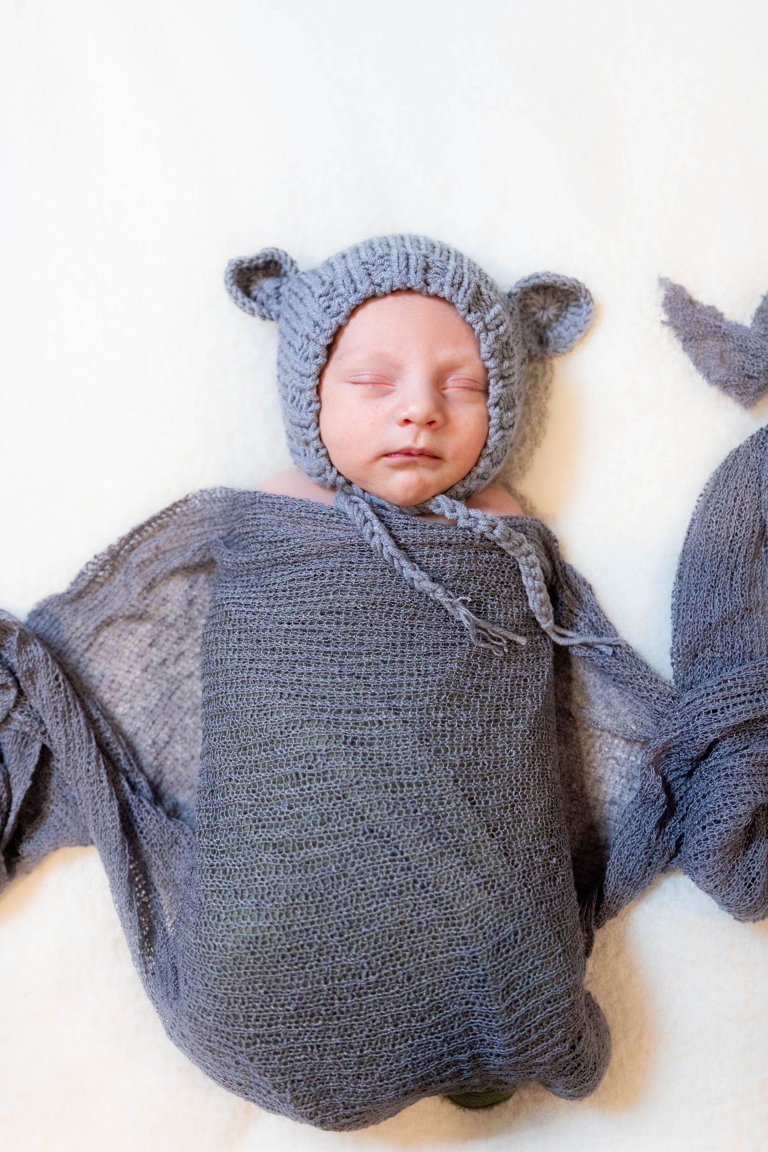 A sleeping baby swaddled in a cozy gray knit blanket with a matching mouse ear hat lies peacefully against a white backdrop.