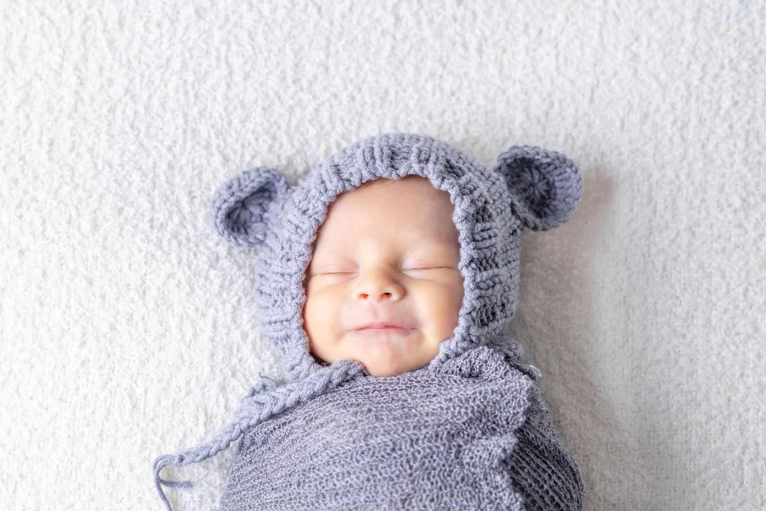Baby wearing cozy gray knit bear hat and wrap lying peacefully on white background.