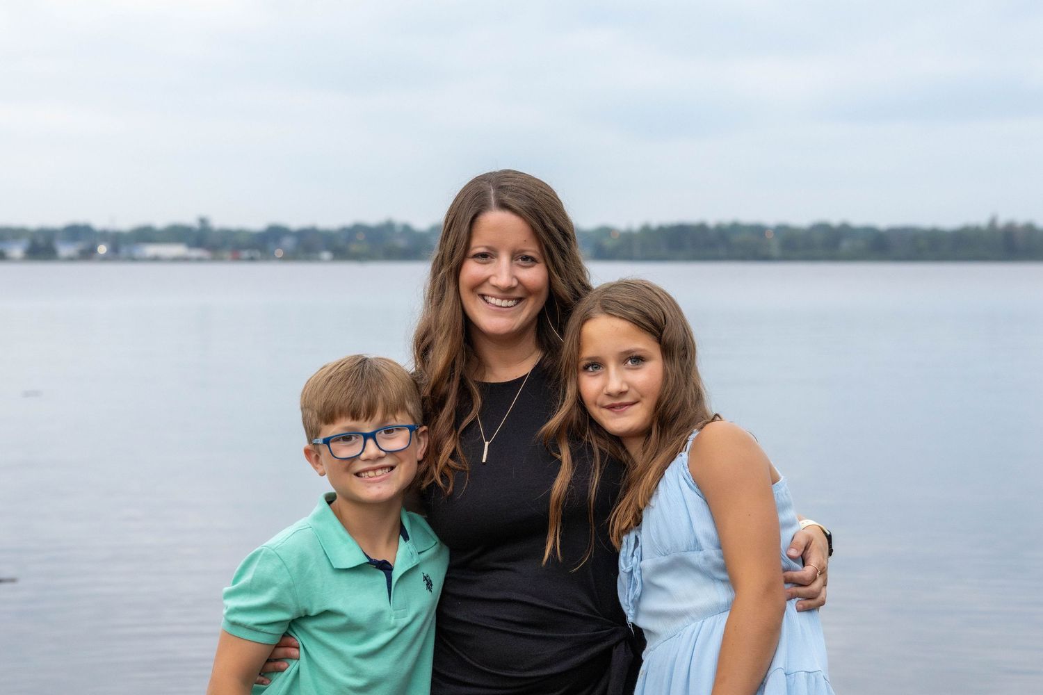 Three people pose together while standing along a waterfront with a calm body of water and distant shoreline in background.