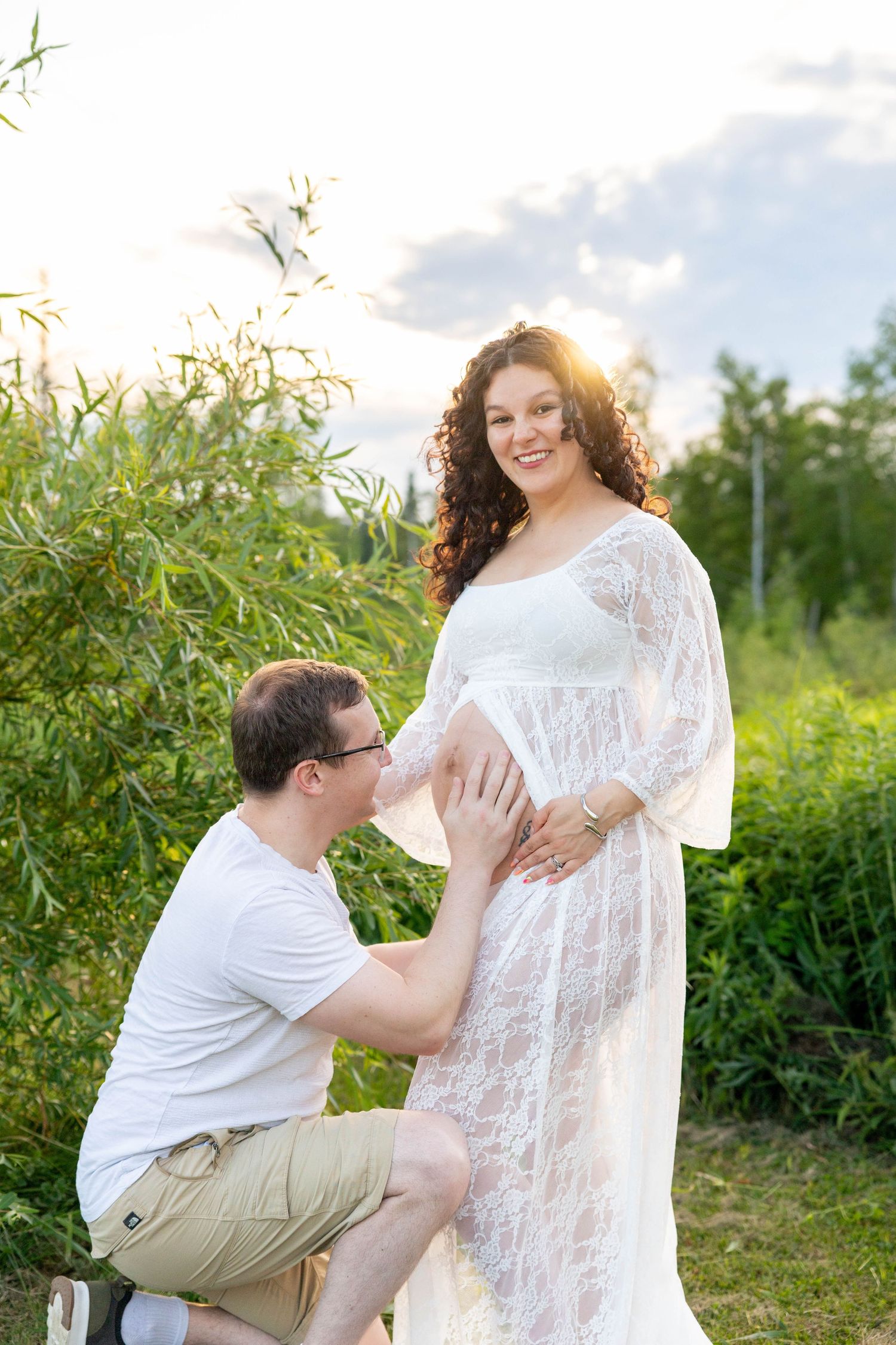 A man kneels touching a pregnant woman's belly outdoors at golden hour, she wears a white lace maternity gown.