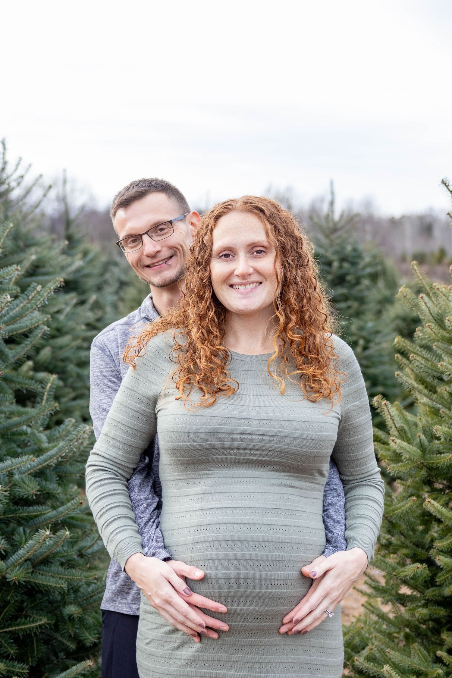 A couple poses together at a Christmas tree farm as they celebrate a pregnancy with hands on the baby bump.