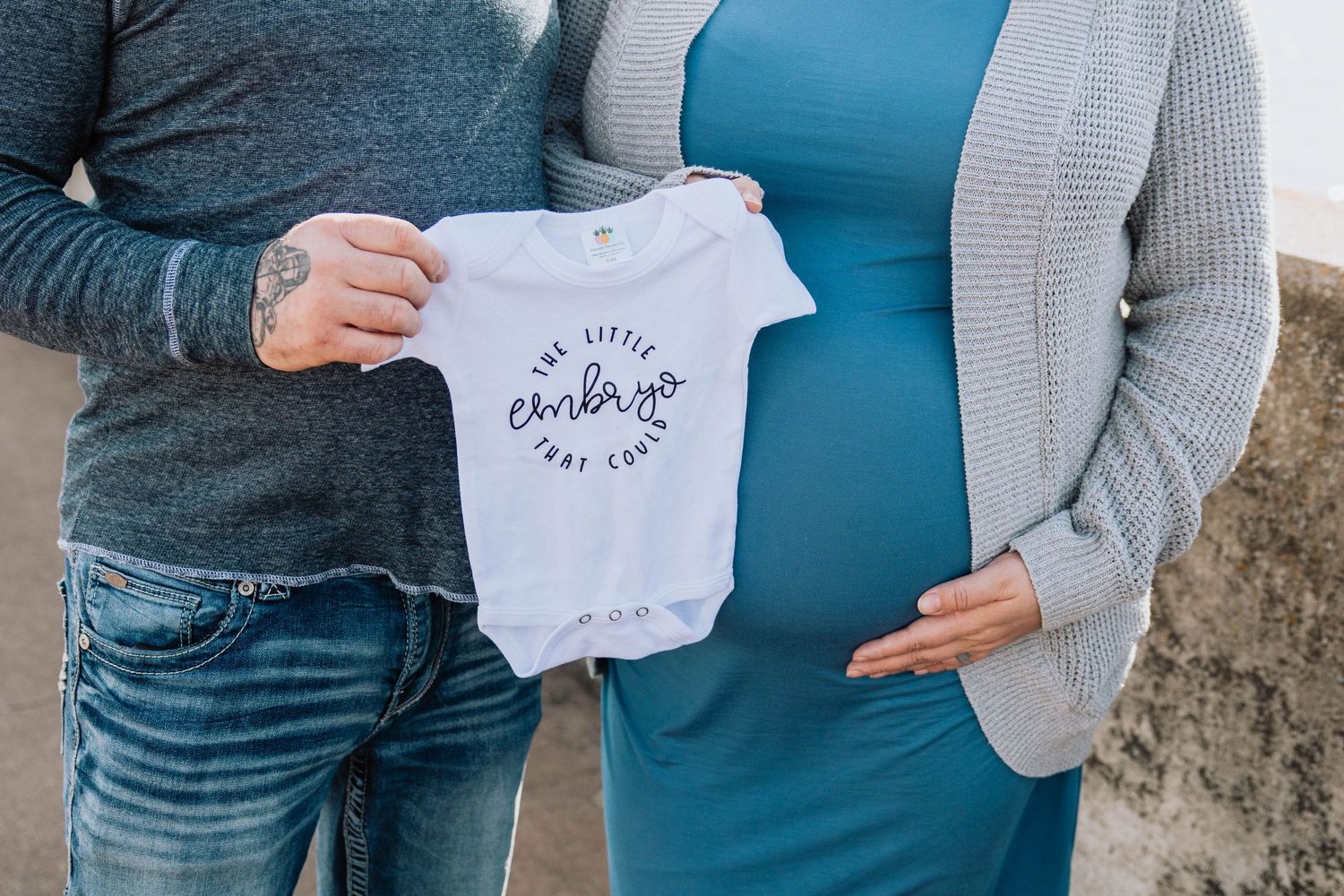A couple holding up a small white baby onesie against a pregnant belly in casual clothing.