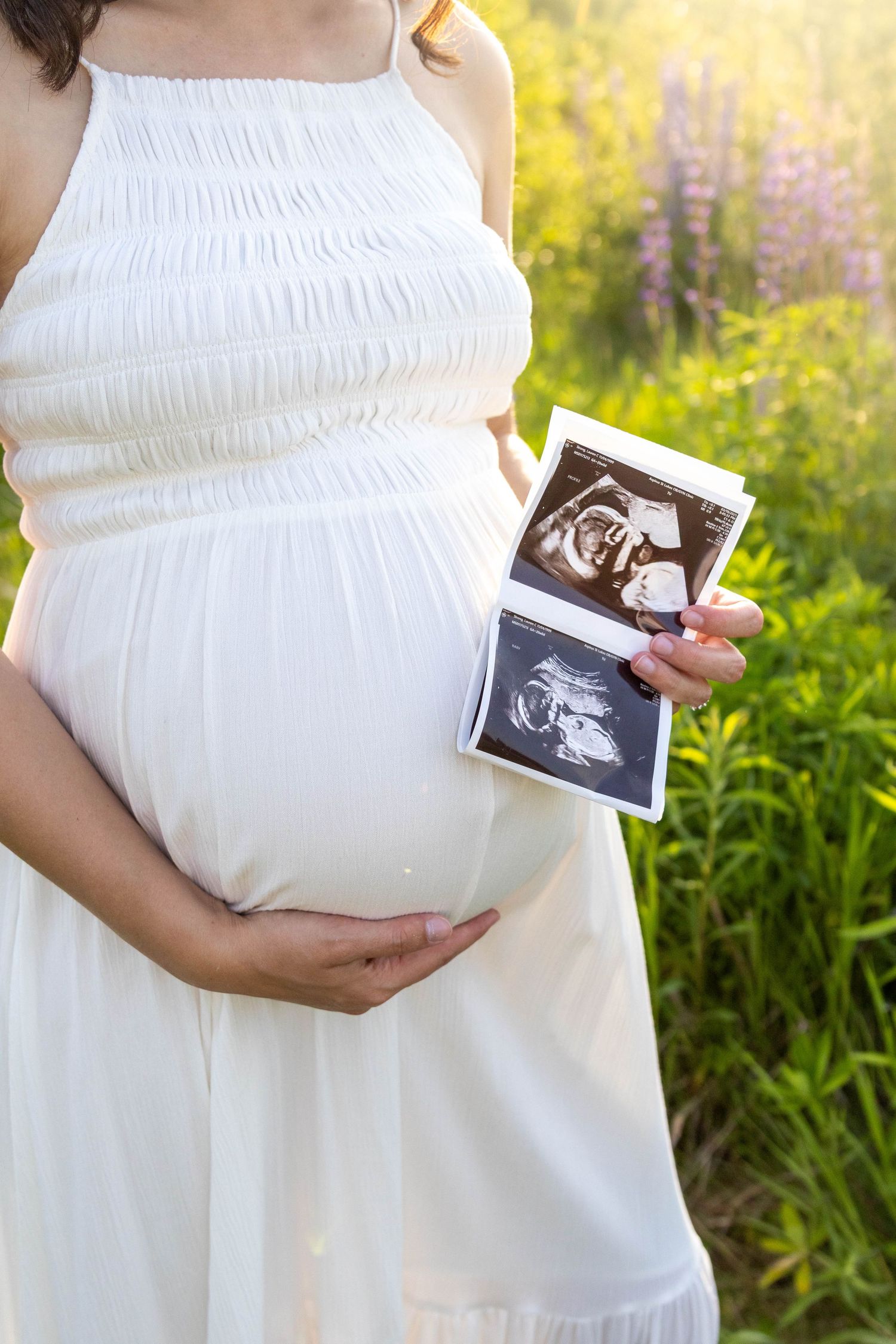 Person in white dress shows ultrasound photos while cradling pregnant belly in a garden with lupine flowers.