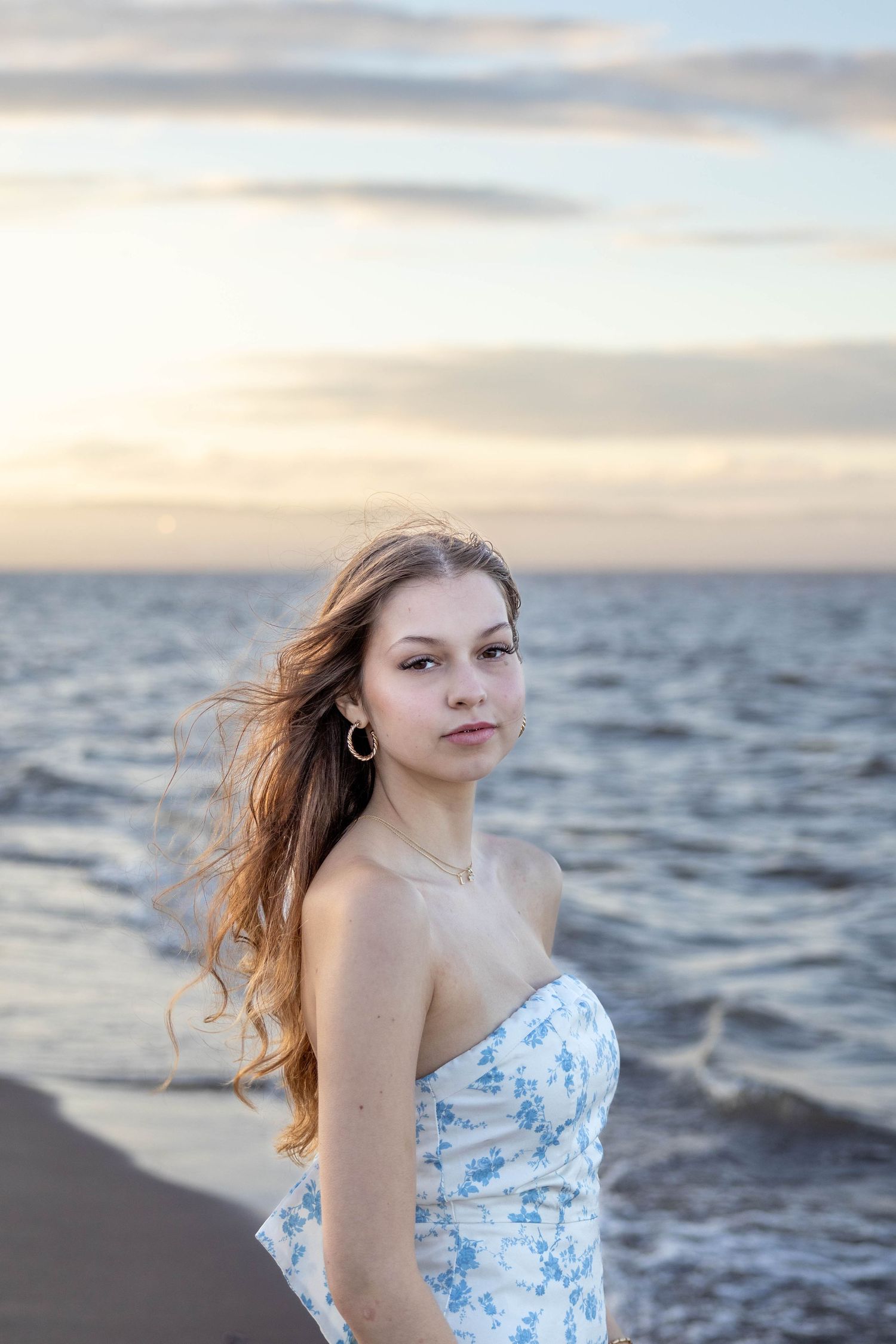 A person in a blue floral dress poses at sunset on a beach with windswept hair near the ocean.