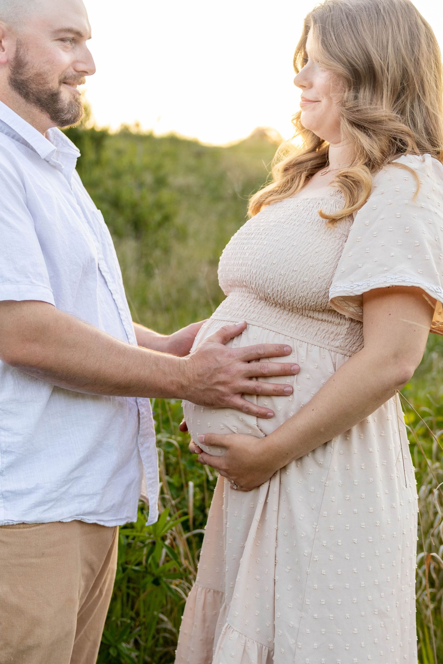 A couple in white and beige clothing embrace during a sunset maternity photoshoot in a grassy field.