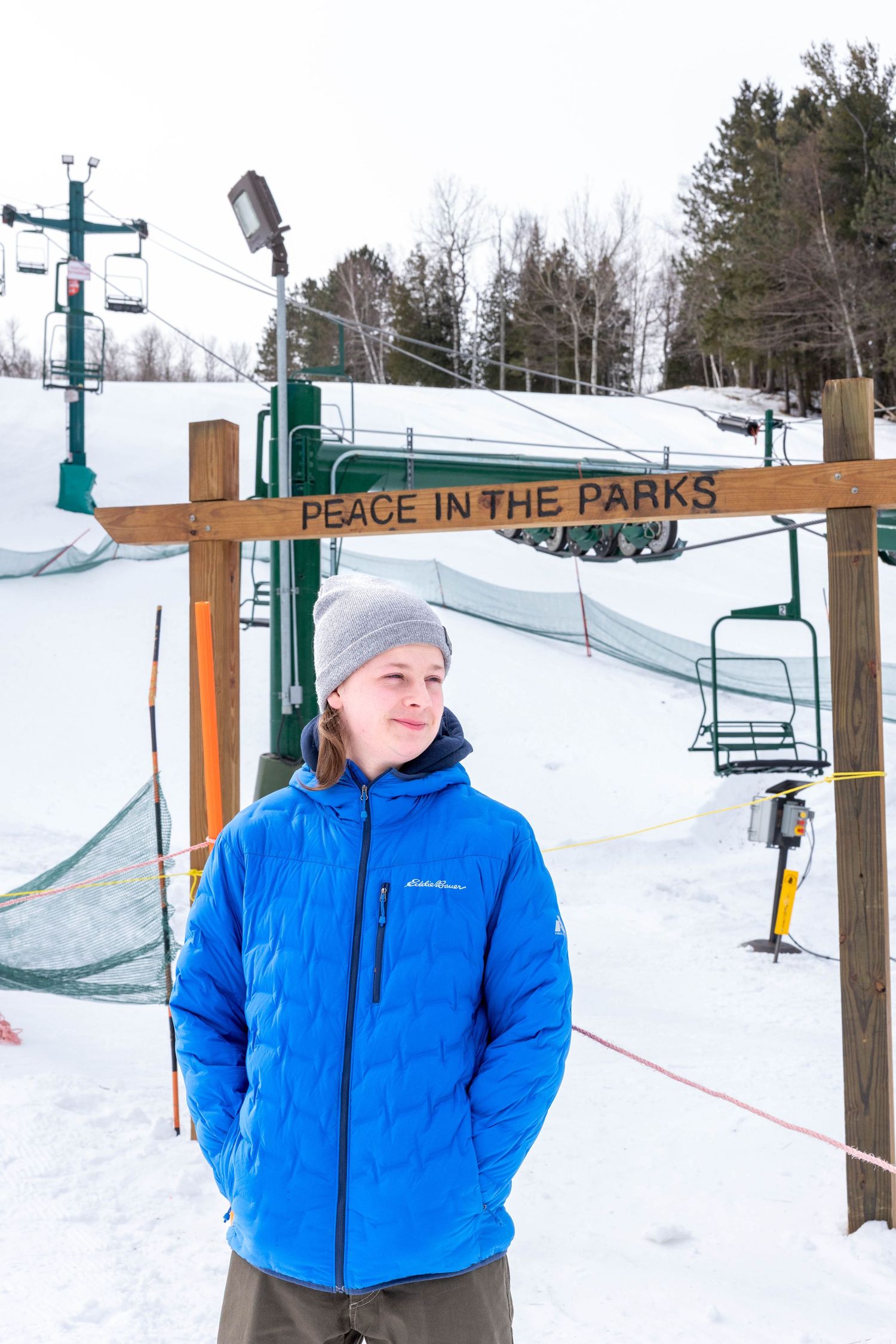 Young person in blue Eddie Bauer jacket stands near Peace in the Parks ski resort sign on snowy winter day.