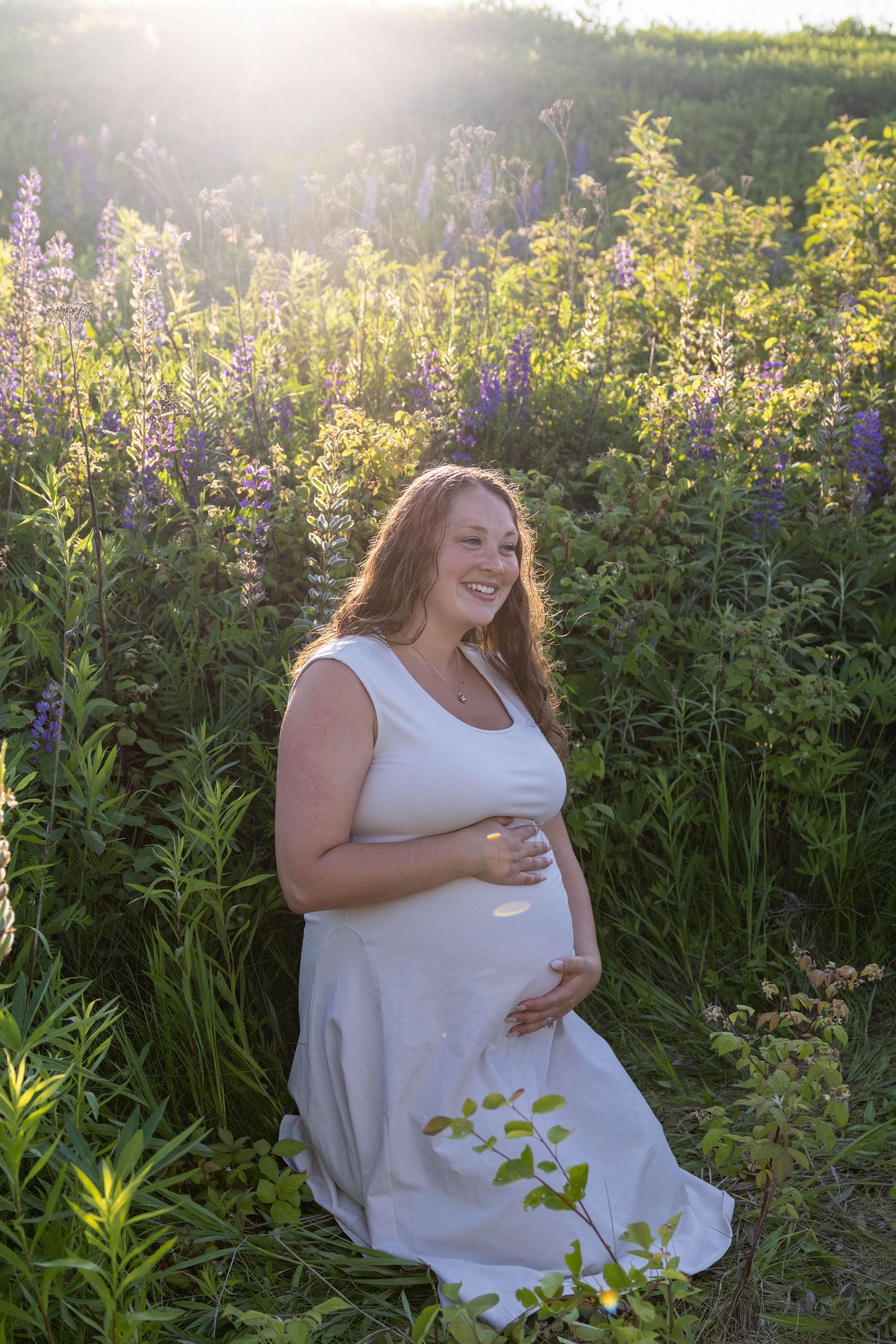 A white dress-clad woman sits in a sun-drenched meadow filled with blooming purple lupines on a summer evening.