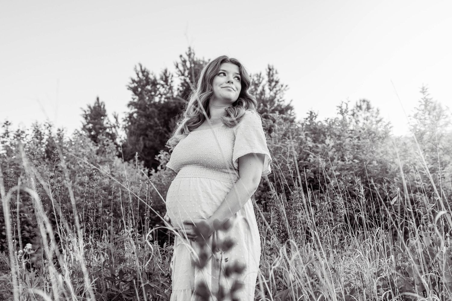 Black and white maternity photo showing expecting mother standing in a field of tall grass during golden hour.