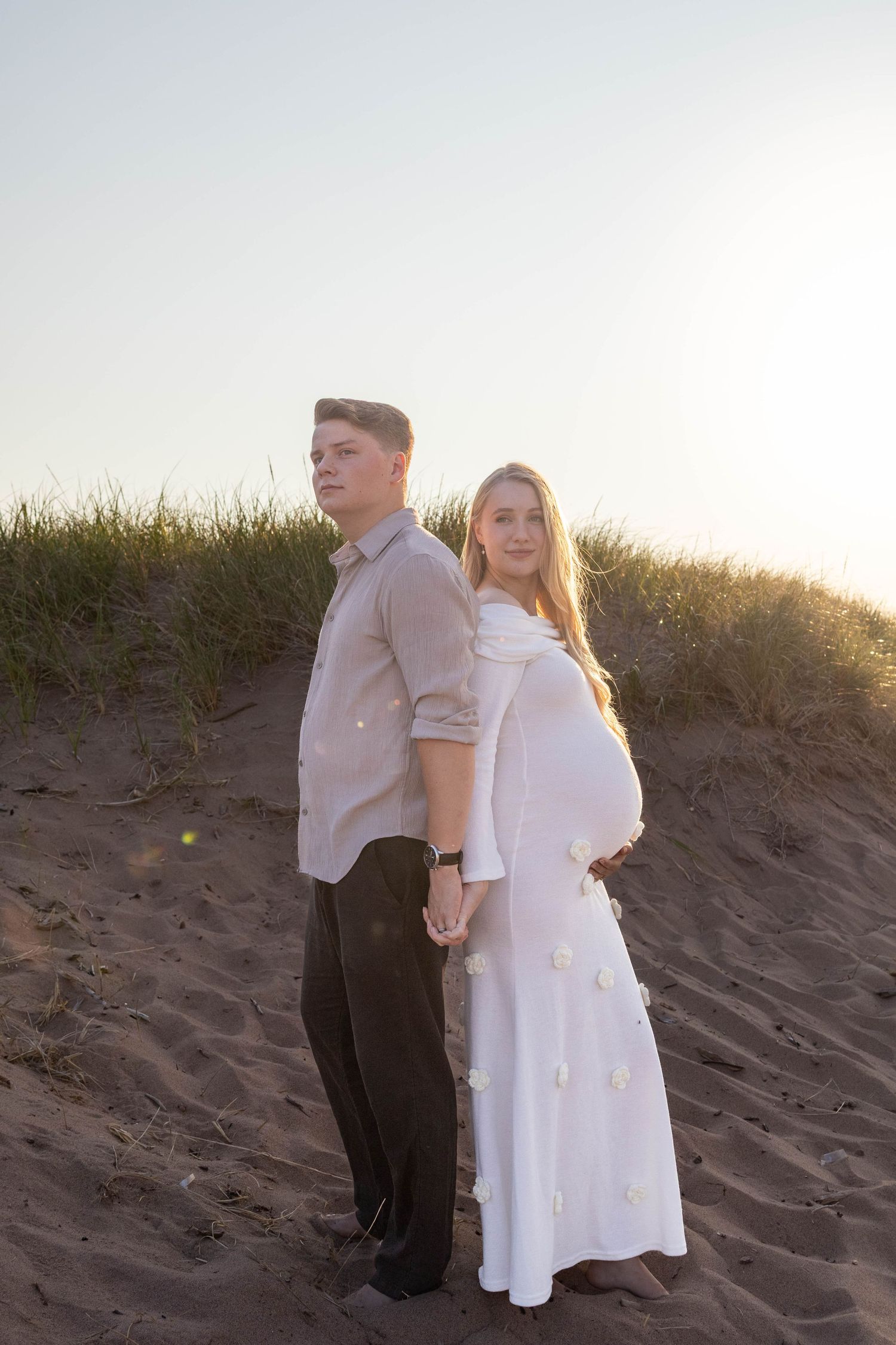 A couple stands together on sandy dunes during a beach photoshoot at sunset wearing formal attire.