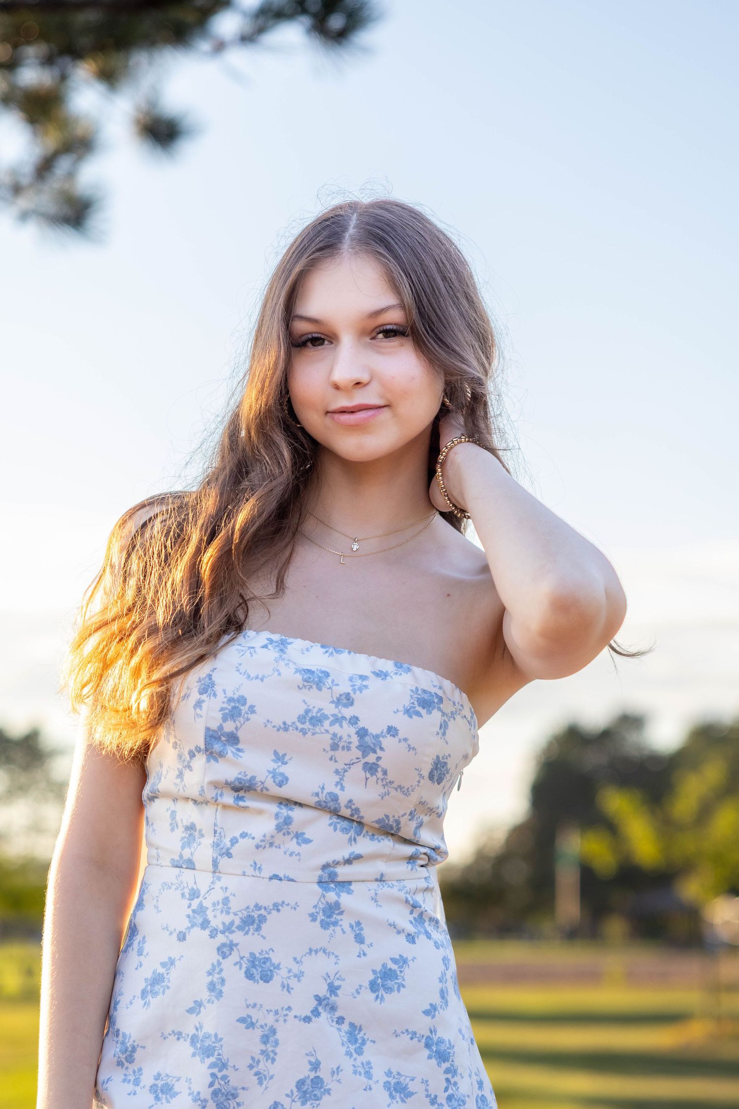 Portrait of someone wearing a strapless white and blue floral dress standing outdoors in warm sunset lighting.