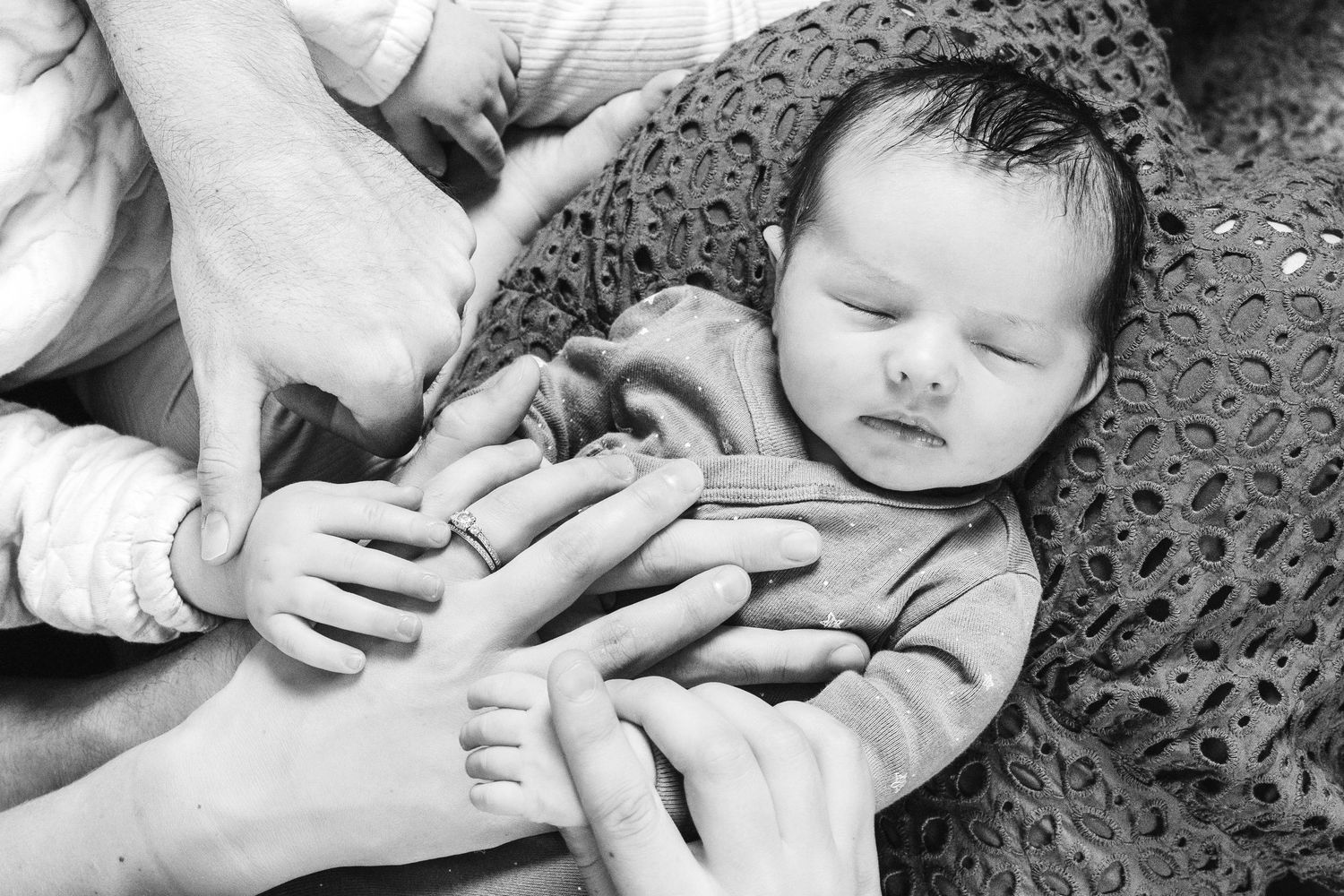 A sleeping baby rests on a textured surface while hands gently touch their tiny fingers in black and white.