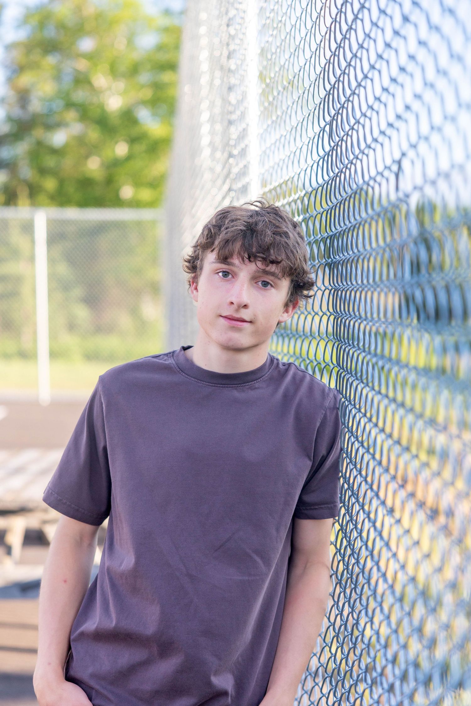 Teen boy in gray t-shirt leaning against chain-link fence outdoors with green trees in background.