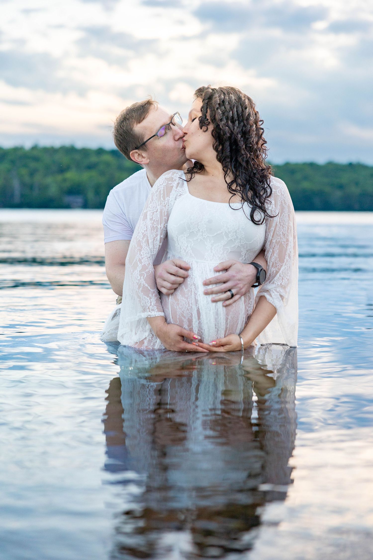 Pregnant couple kissing while standing in a lake, man embracing woman's baby bump, cloudy sky and trees in background.