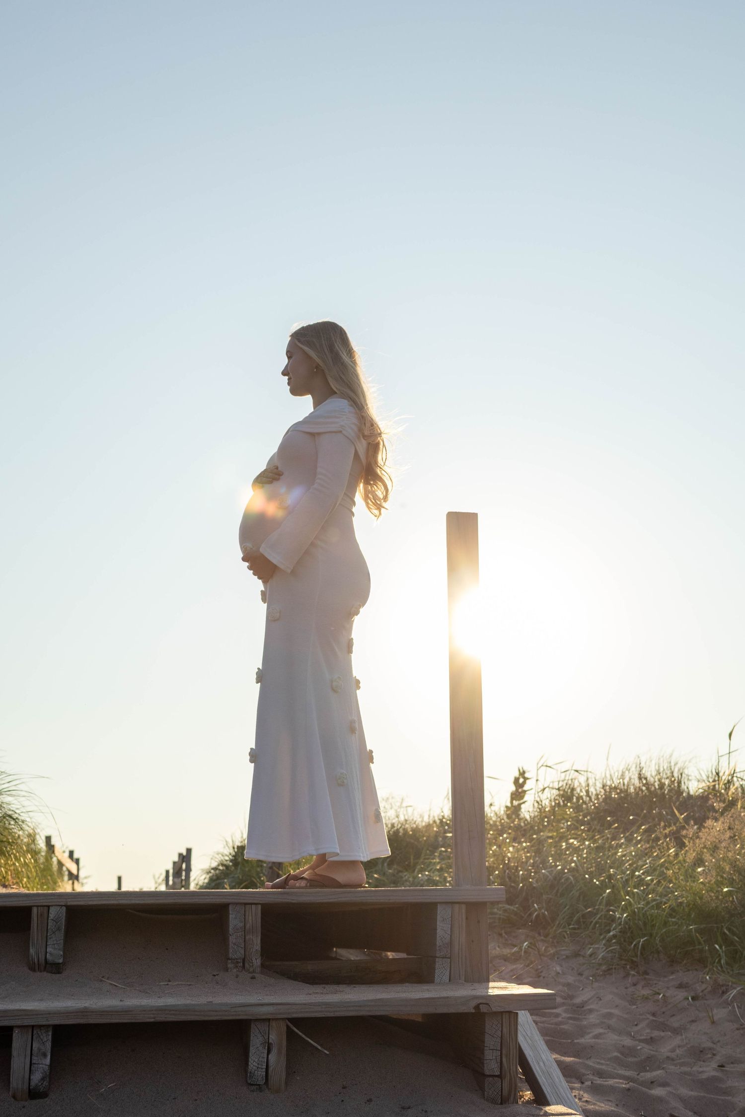 Silhouette of pregnant woman in white dress standing on wooden boardwalk at beach during golden sunset.