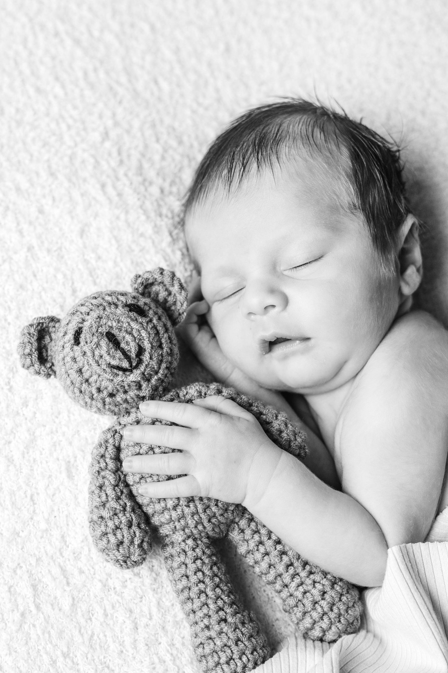 Black and white image of sleeping newborn cuddling knit teddy bear.