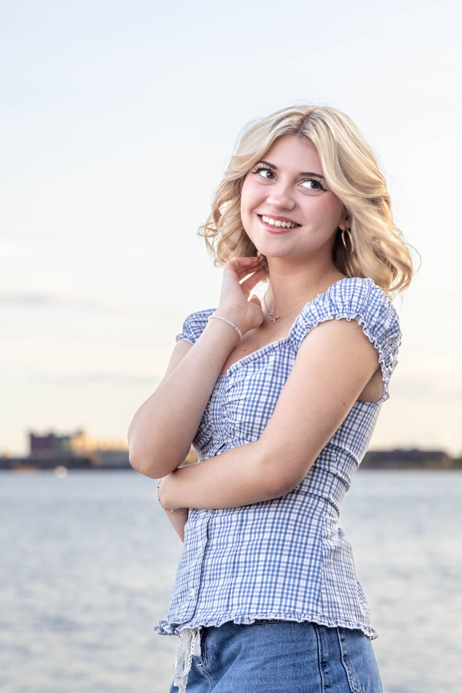 Young woman in plaid ruffle blouse and denim smiles while posing outdoors near water at sunset.
