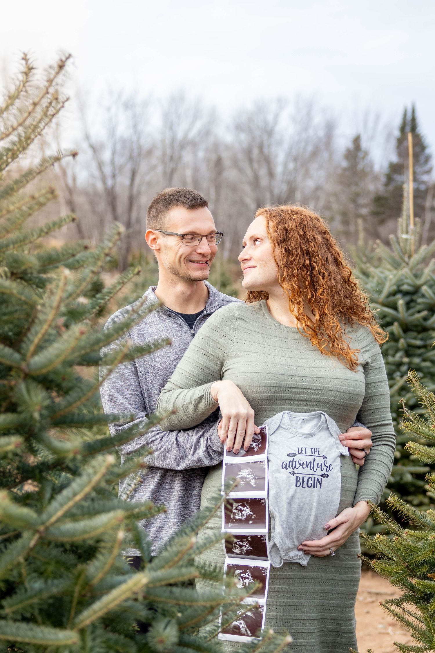 A couple embraces among evergreen trees while holding ultrasound photos during a pregnancy announcement photoshoot.