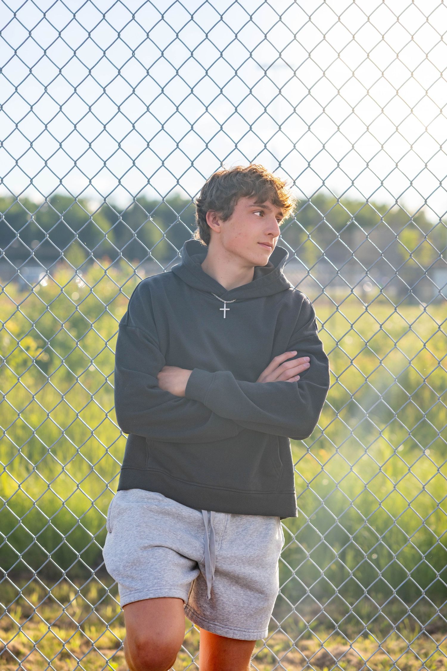 Teen boy in gray hoodie and shorts leaning against chain-link fence outdoors in natural sunlight.