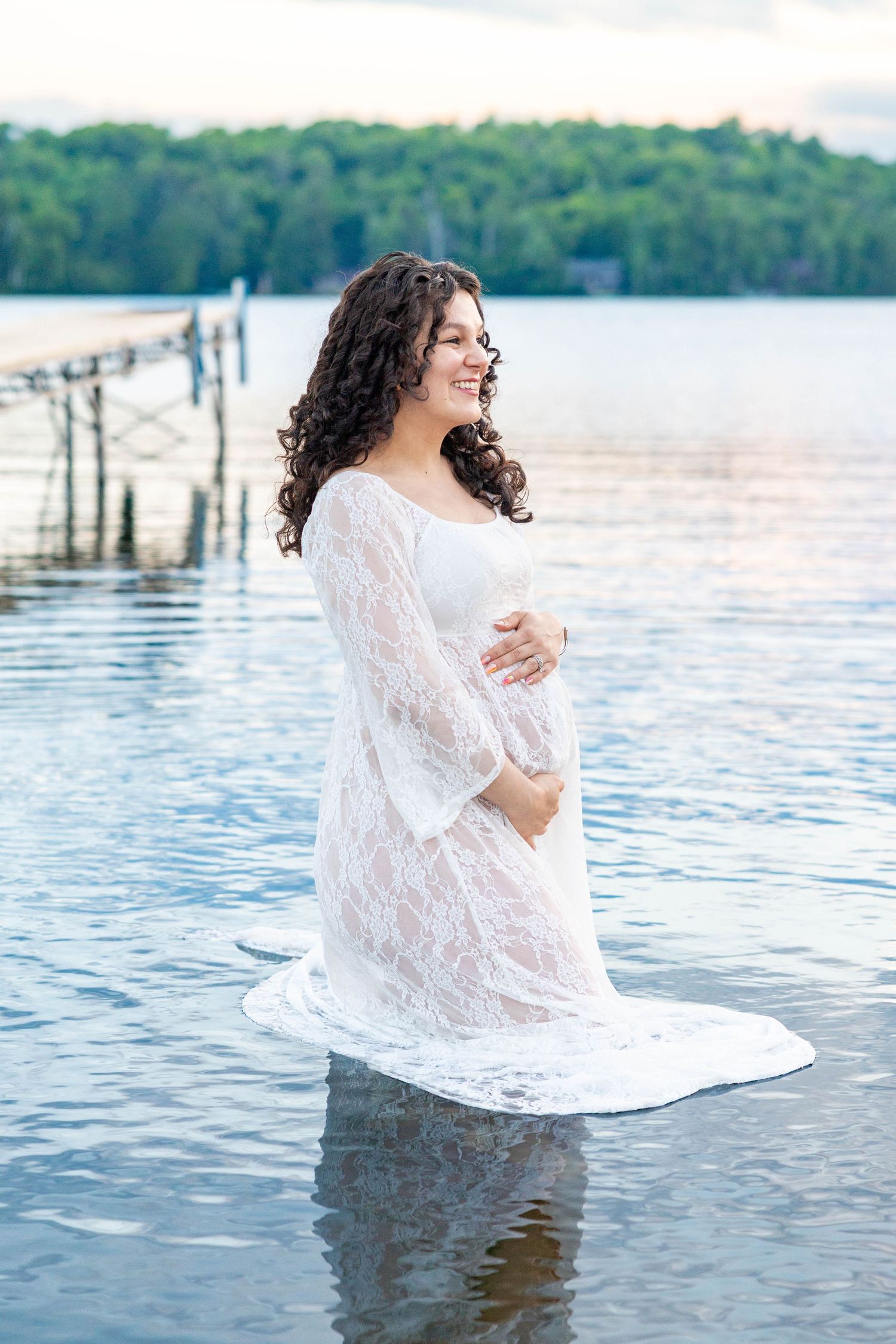 Pregnant woman in white lace dress standing in calm lake water, smiling serenely with a dock and trees in background.