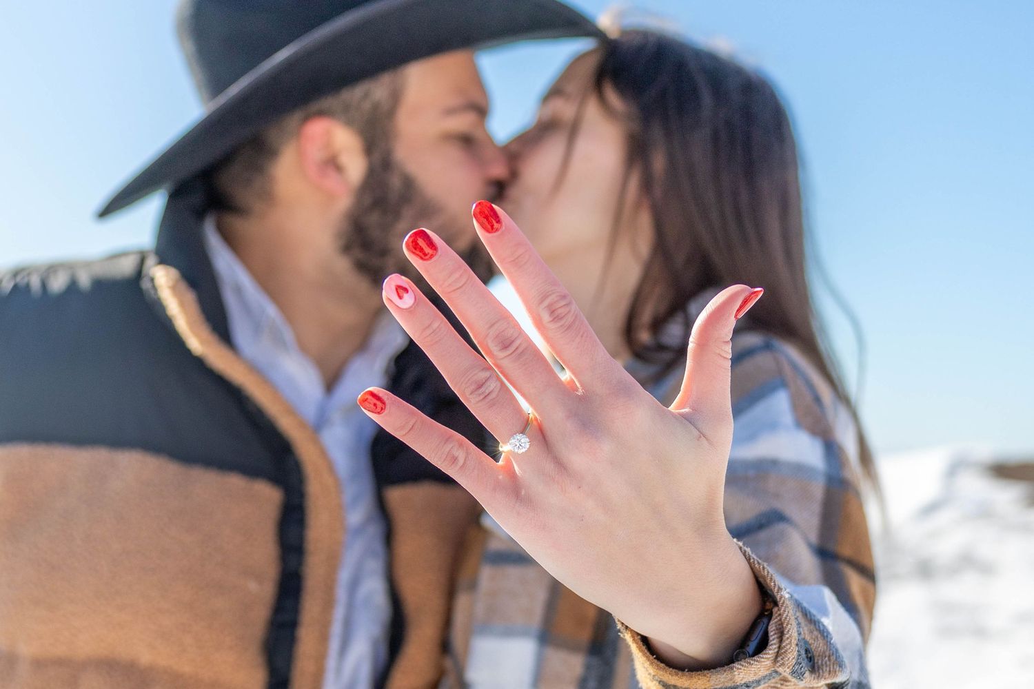 Two people share a romantic kiss against a snowy winter mountain backdrop while holding up their hands showing engagement rings.