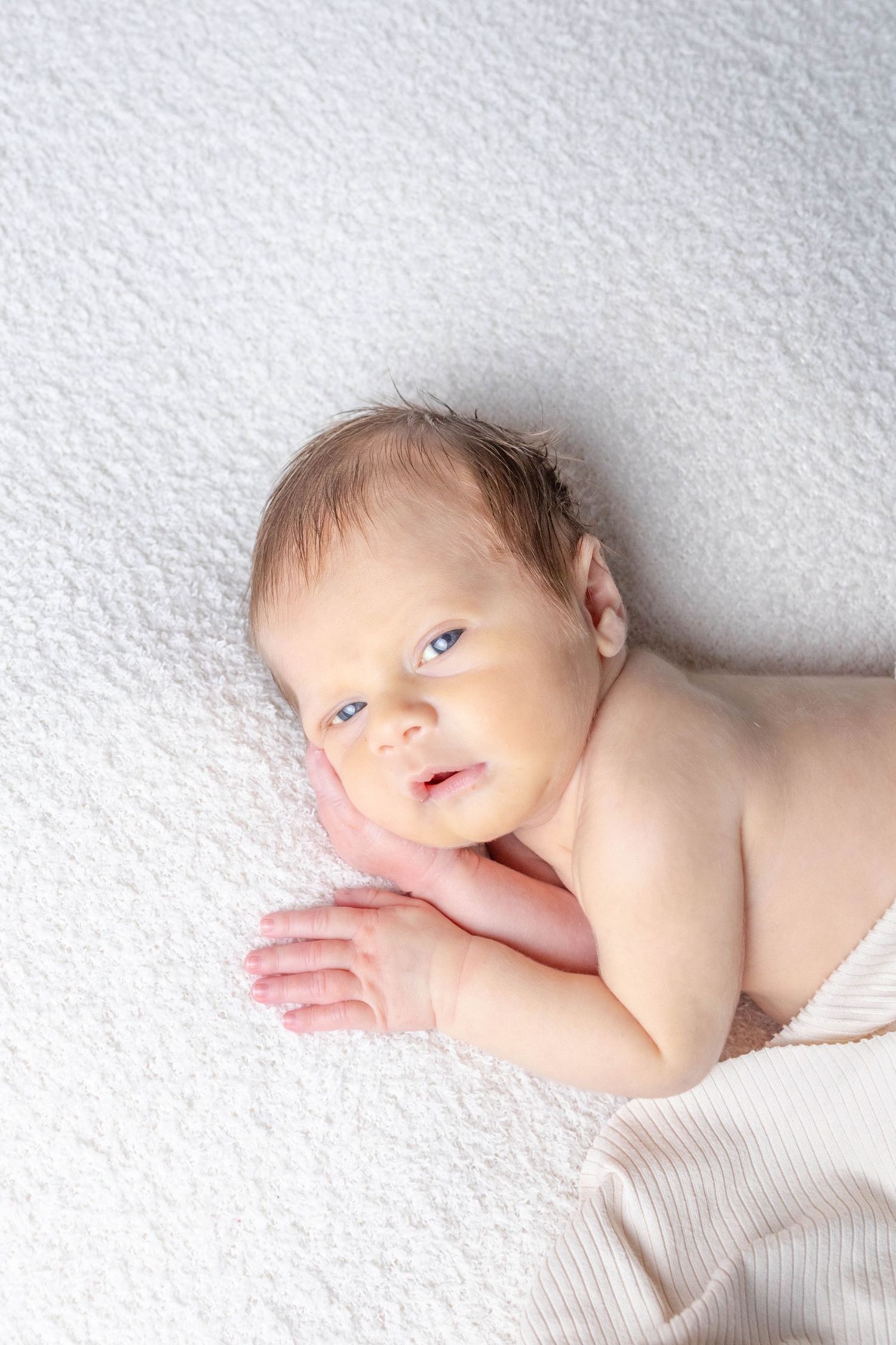 Alert newborn baby lying on white textured background looking at camera.