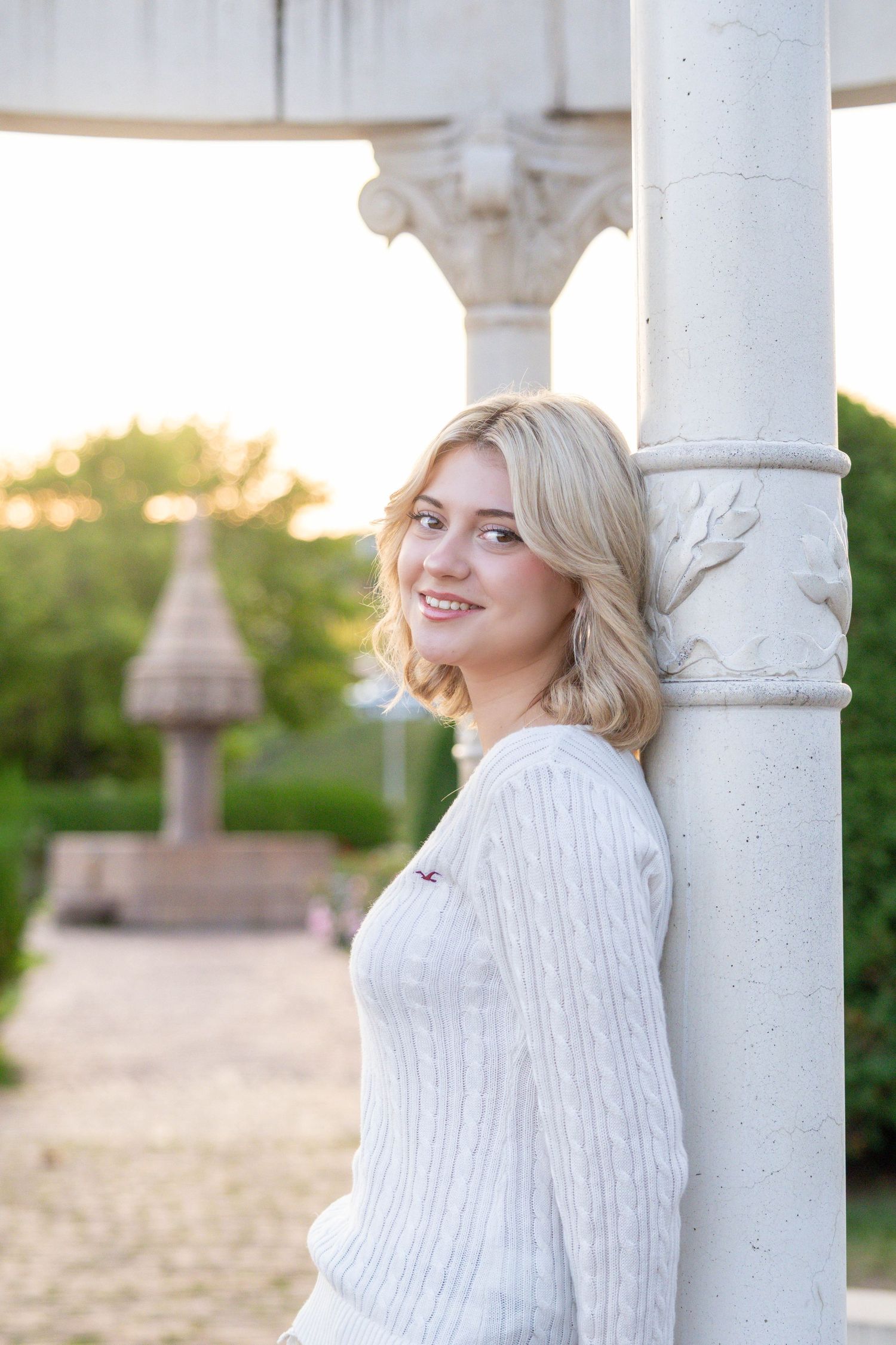 Woman in a white knit sweater poses elegantly against white columns in an outdoor setting during golden hour.