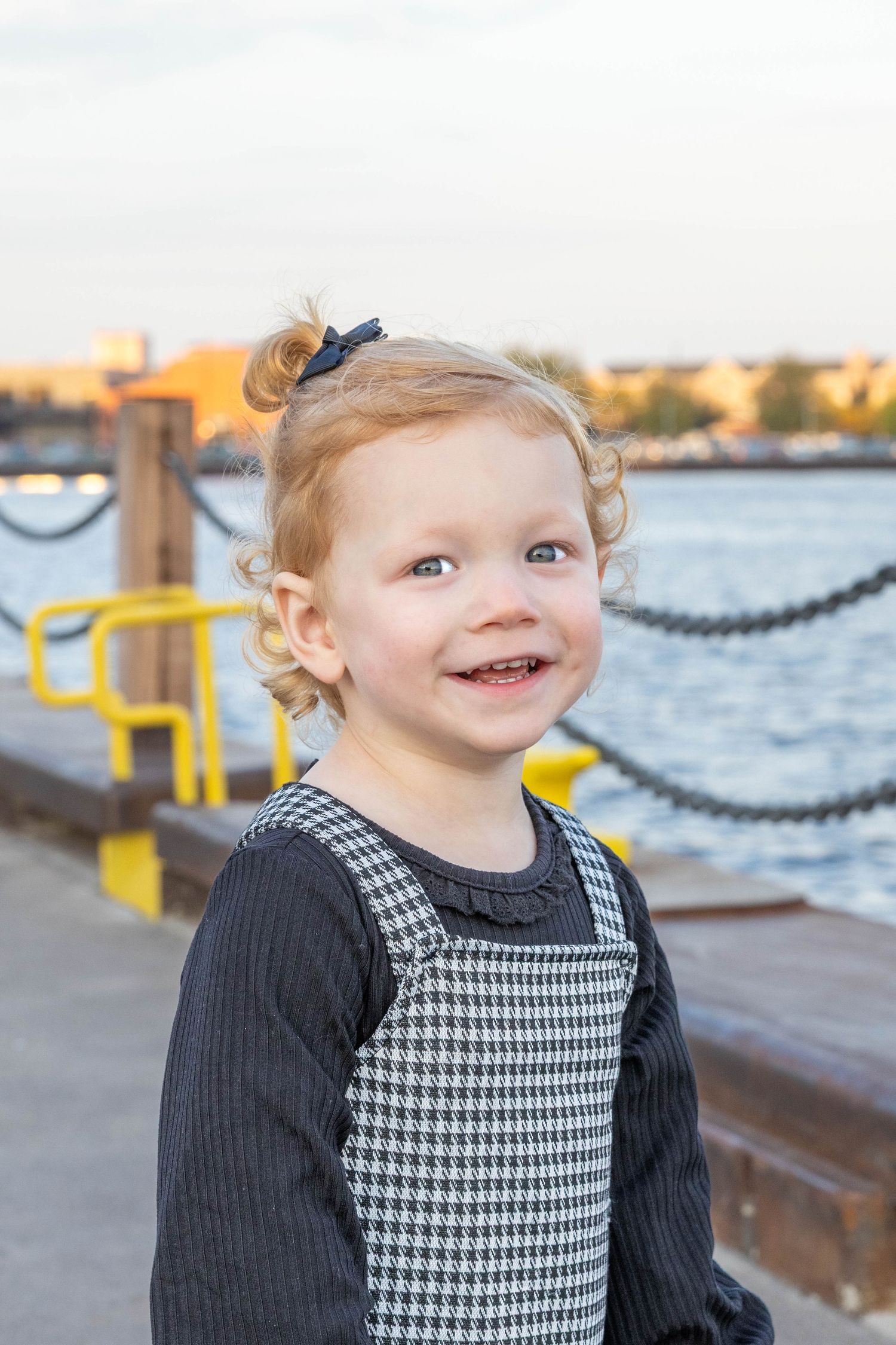 A young child in a black and houndstooth dress smiles while standing near a waterfront railing at sunset.