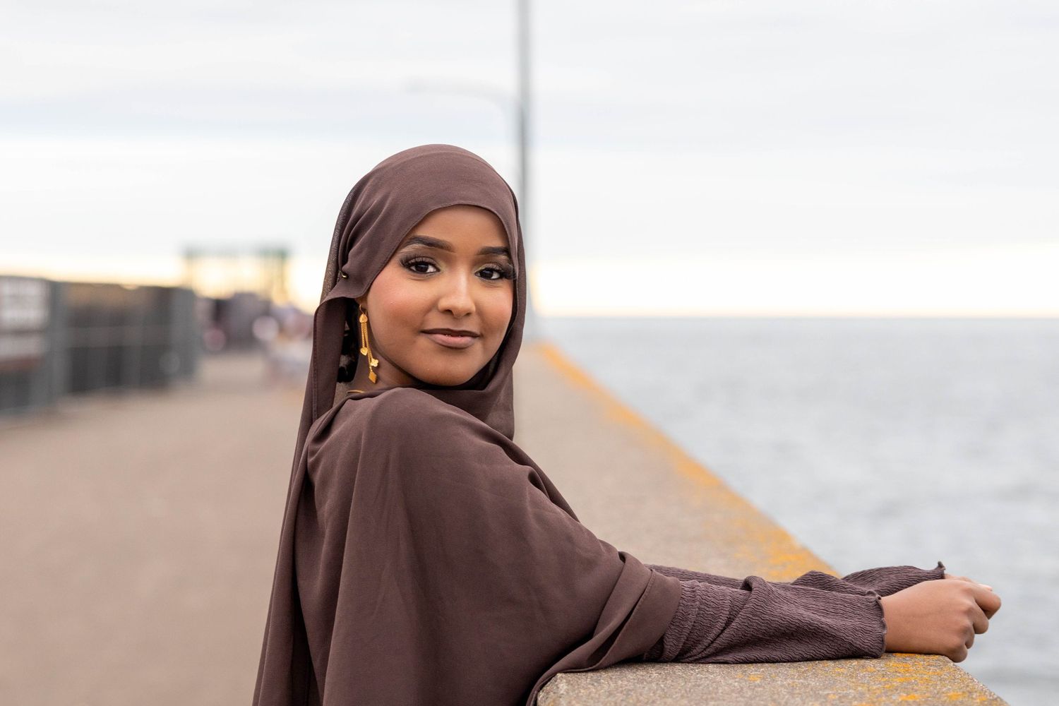 Woman in brown hijab smiles while posing outdoors near a body of water.