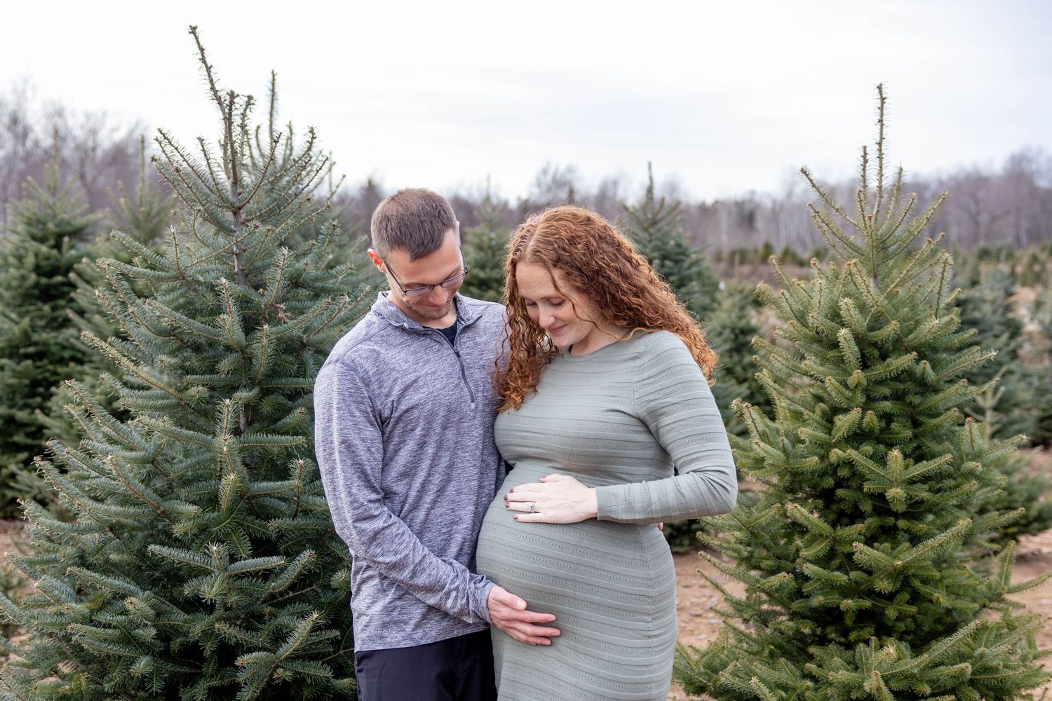 A couple embraces among evergreen trees at a Christmas tree farm during a winter maternity photoshoot.