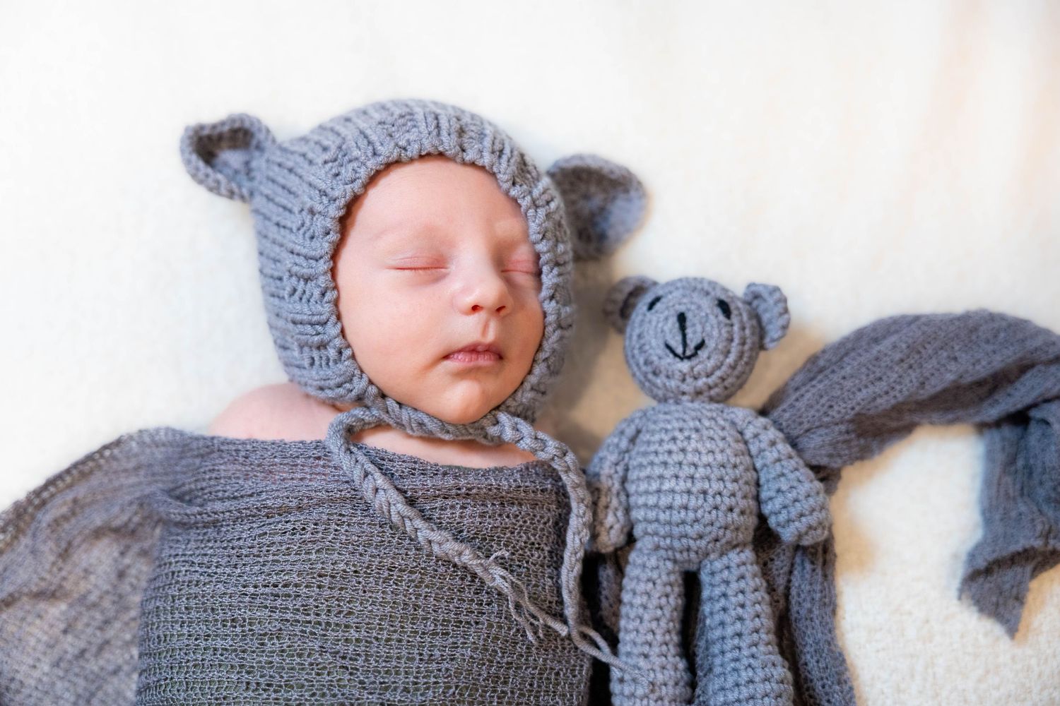 A sleeping baby wearing a gray knit hat rests next to a matching crocheted stuffed elephant toy.