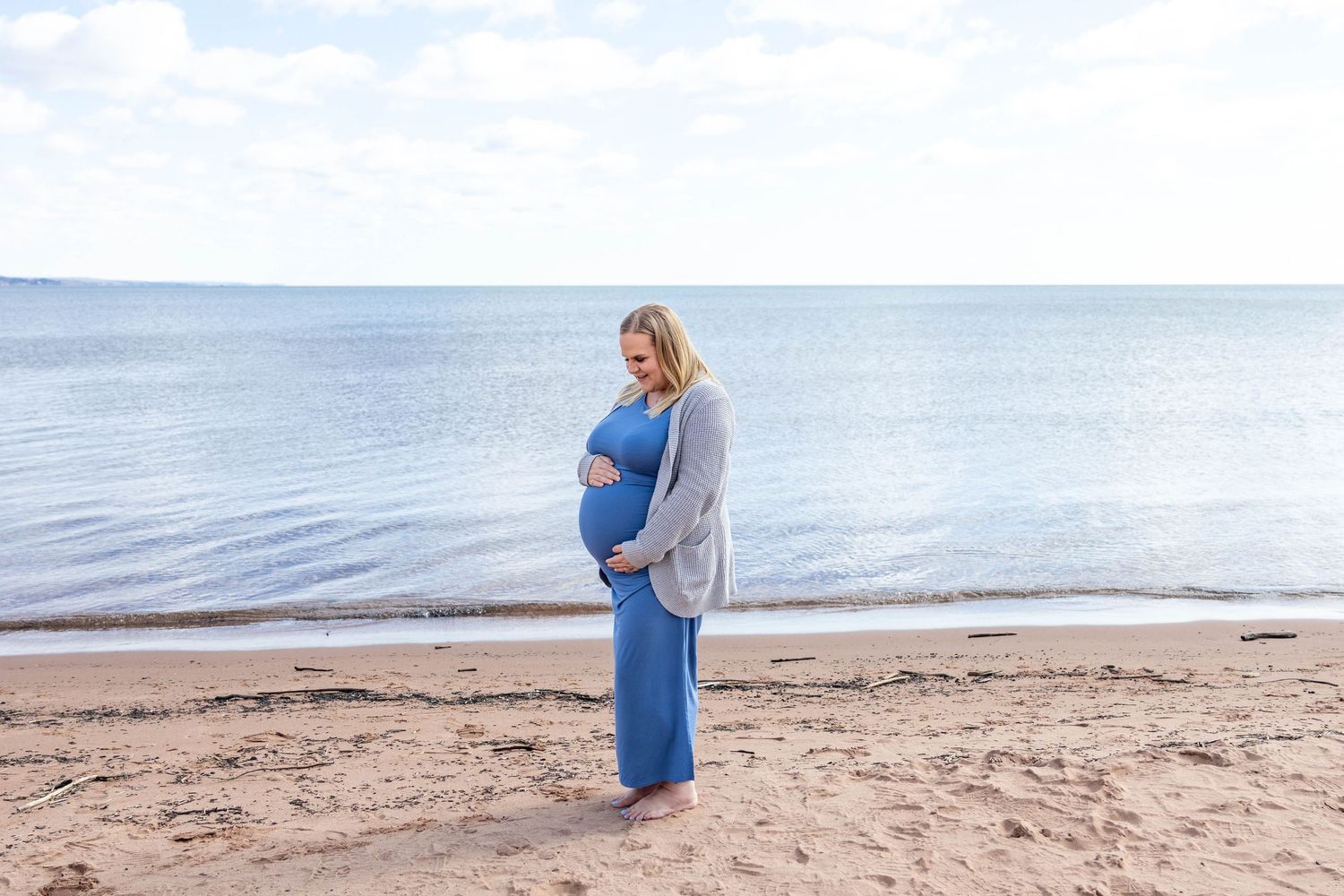 A figure in a flowing blue dress and gray cardigan stands on the beach while cradling their pregnant belly.