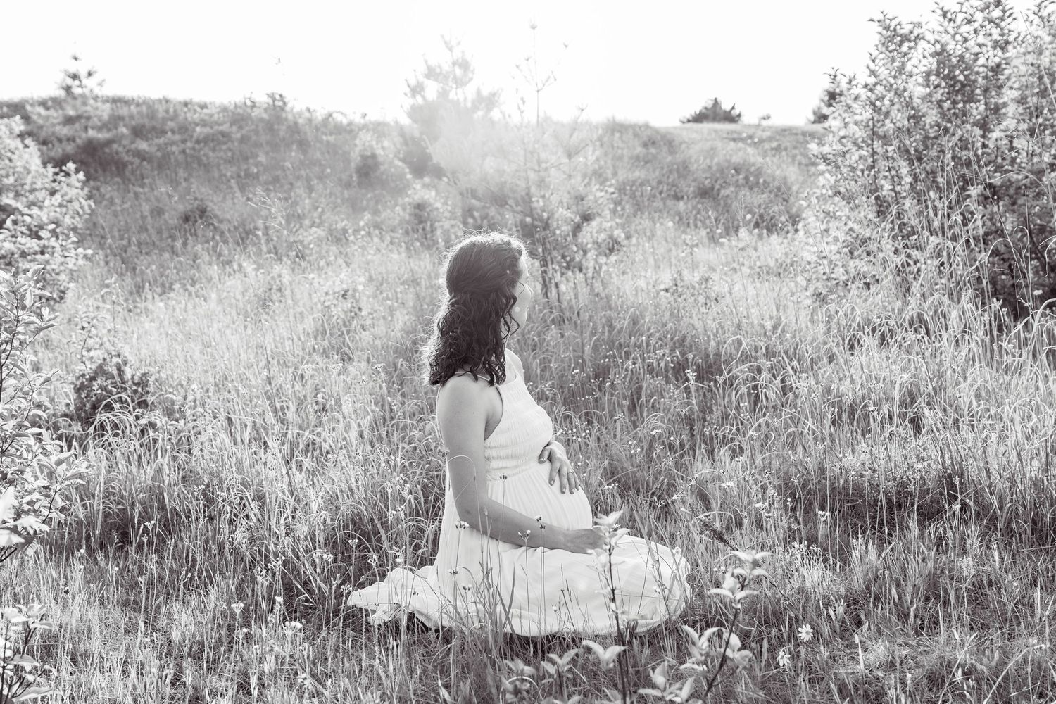 A person in a white dress sits alone in a sunlit meadow surrounded by tall grass, captured in black and white.