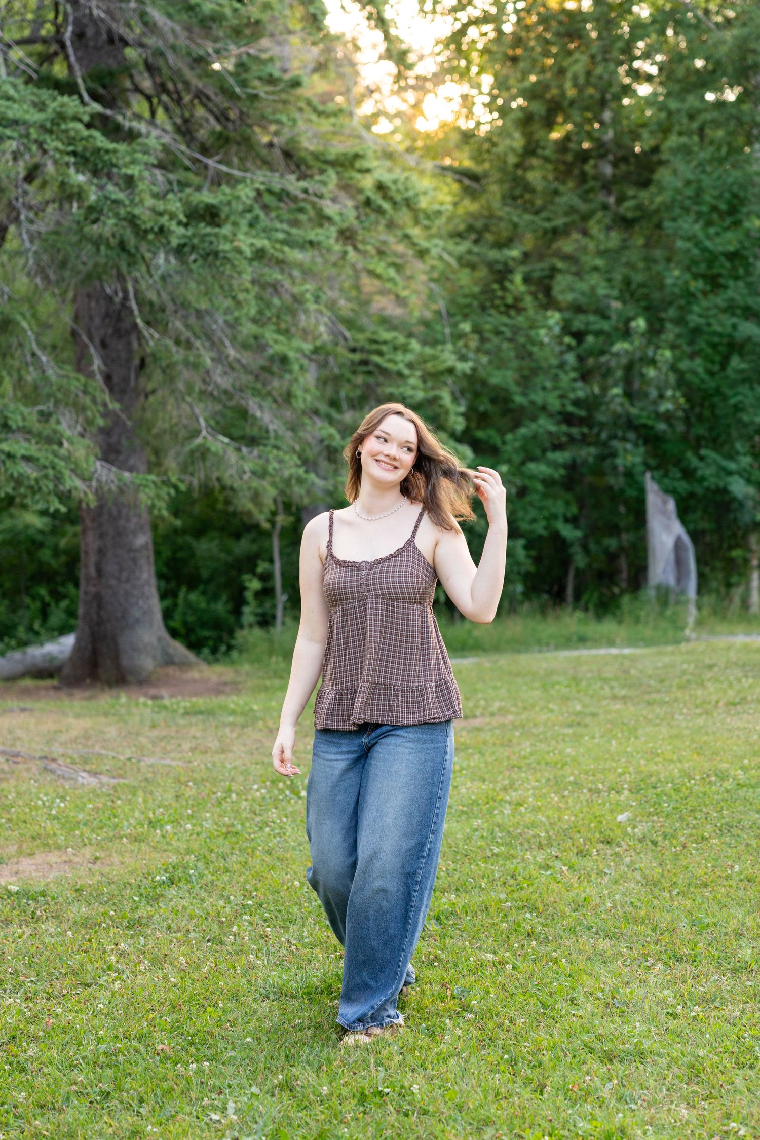 Young woman in brown tank top and jeans smiling while walking on green grass with trees in background.