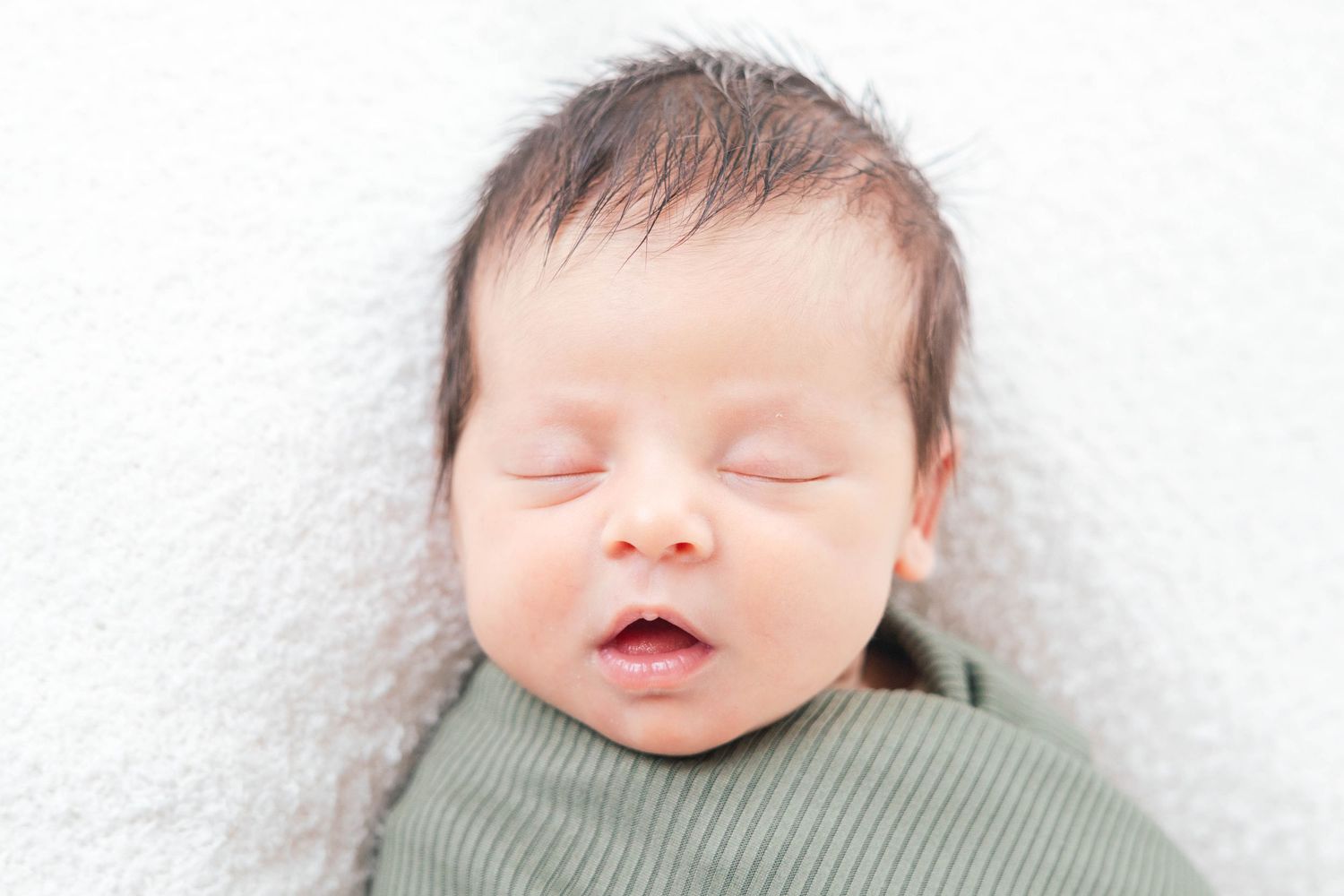 Sleeping newborn in sage green ribbed outfit on white textured background.