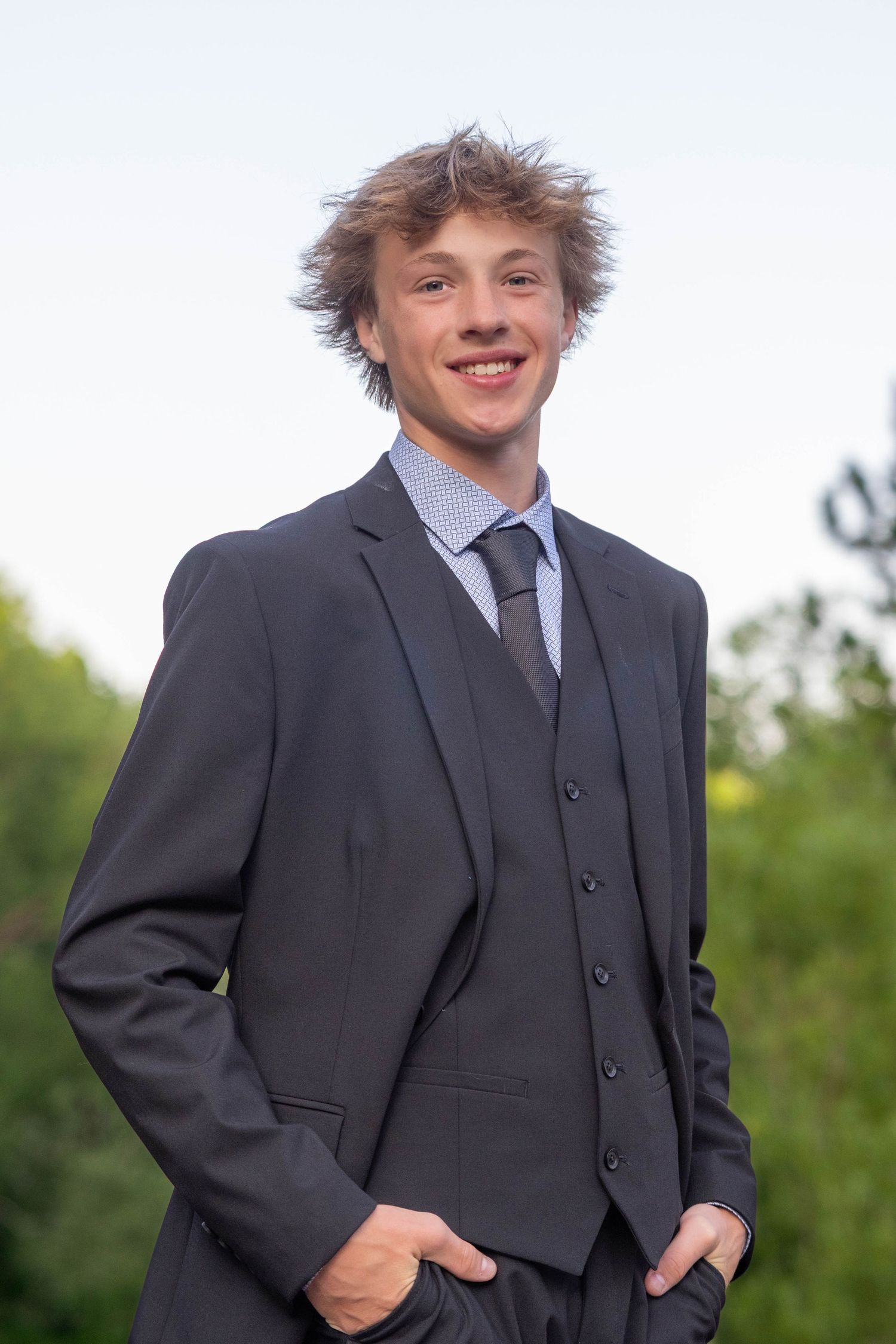 A person in a dark suit and tie poses outdoors against a green foliage background.