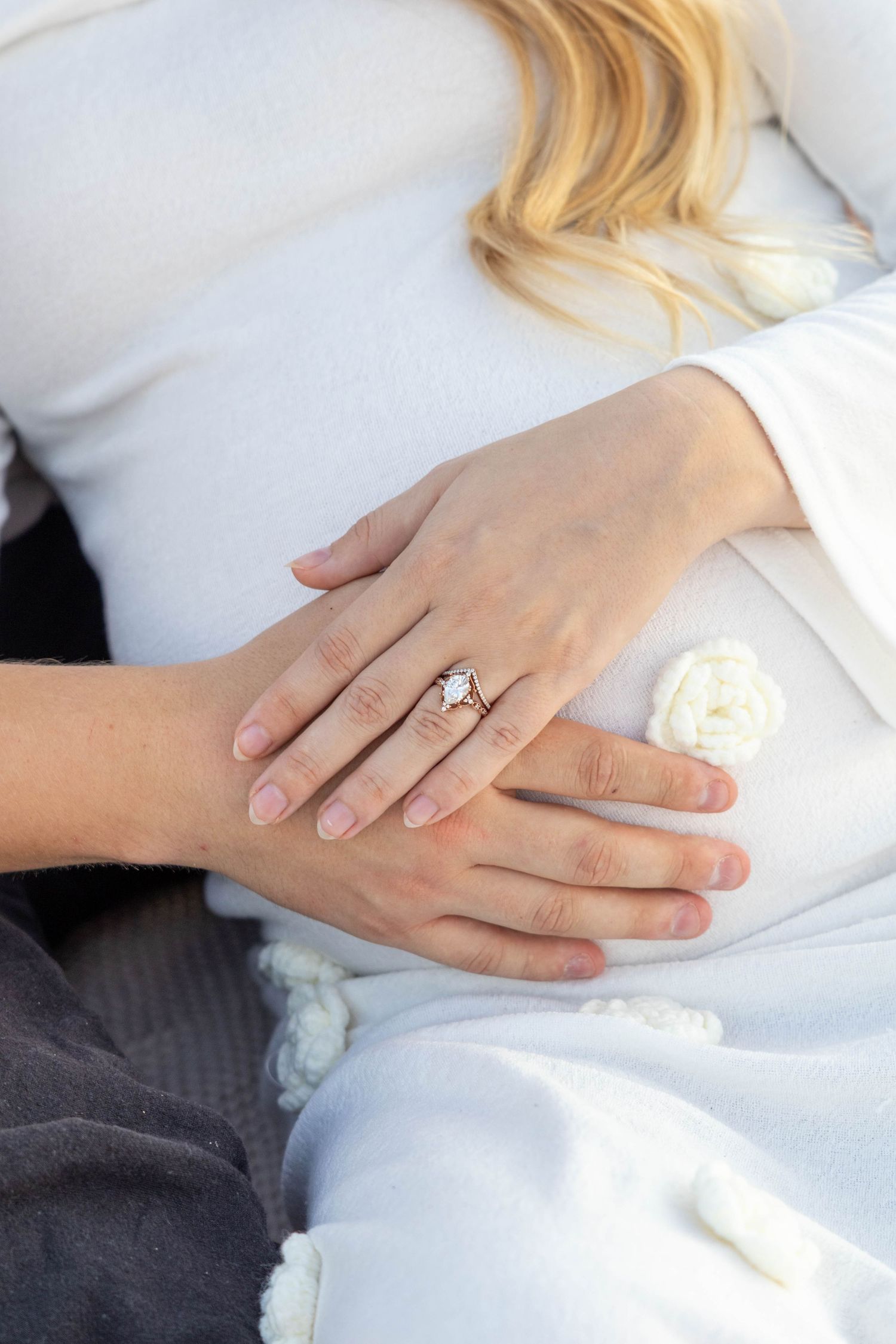 Close-up view of hands wearing a diamond engagement ring on white fabric with soft lighting.