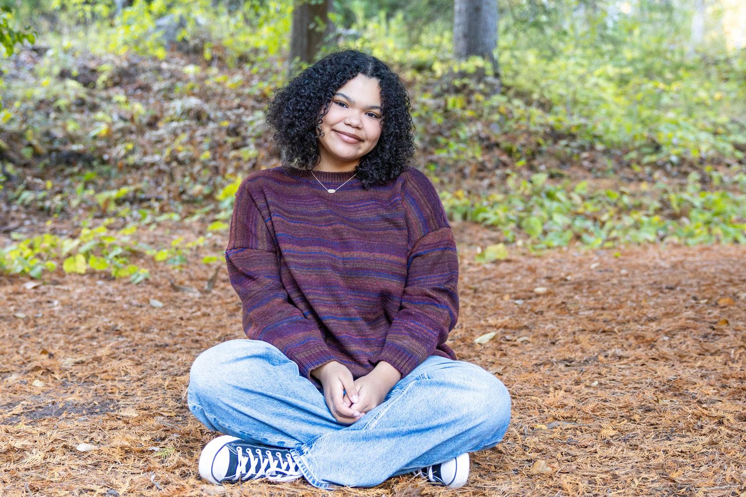 A person in a burgundy sweater and jeans sits cross-legged on fallen autumn leaves in an outdoor setting.