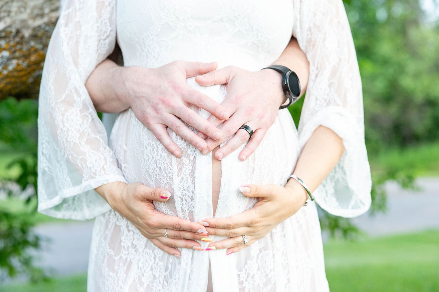 Pregnant woman in white lace dress with partner's hands forming heart shape over baby bump outdoors.