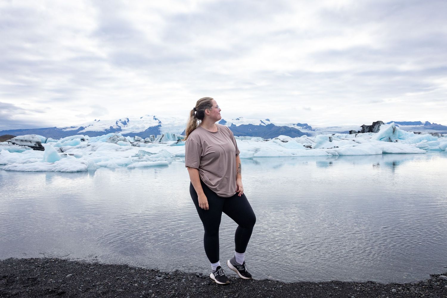 Woman standing on rocky shore with glacial icebergs floating in lagoon under cloudy sky in Iceland.