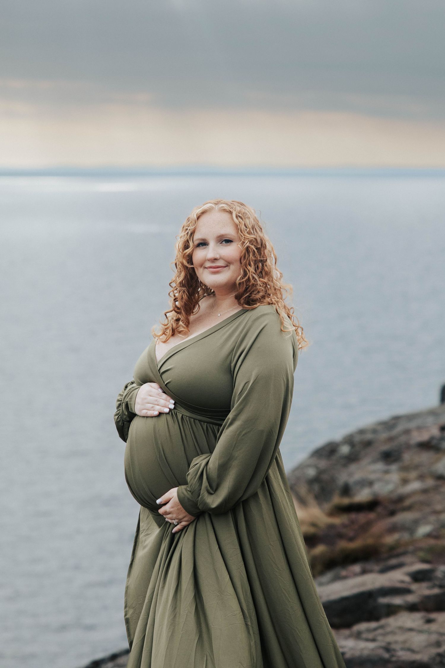 Dramatic maternity portrait of woman in olive dress against moody seascape.