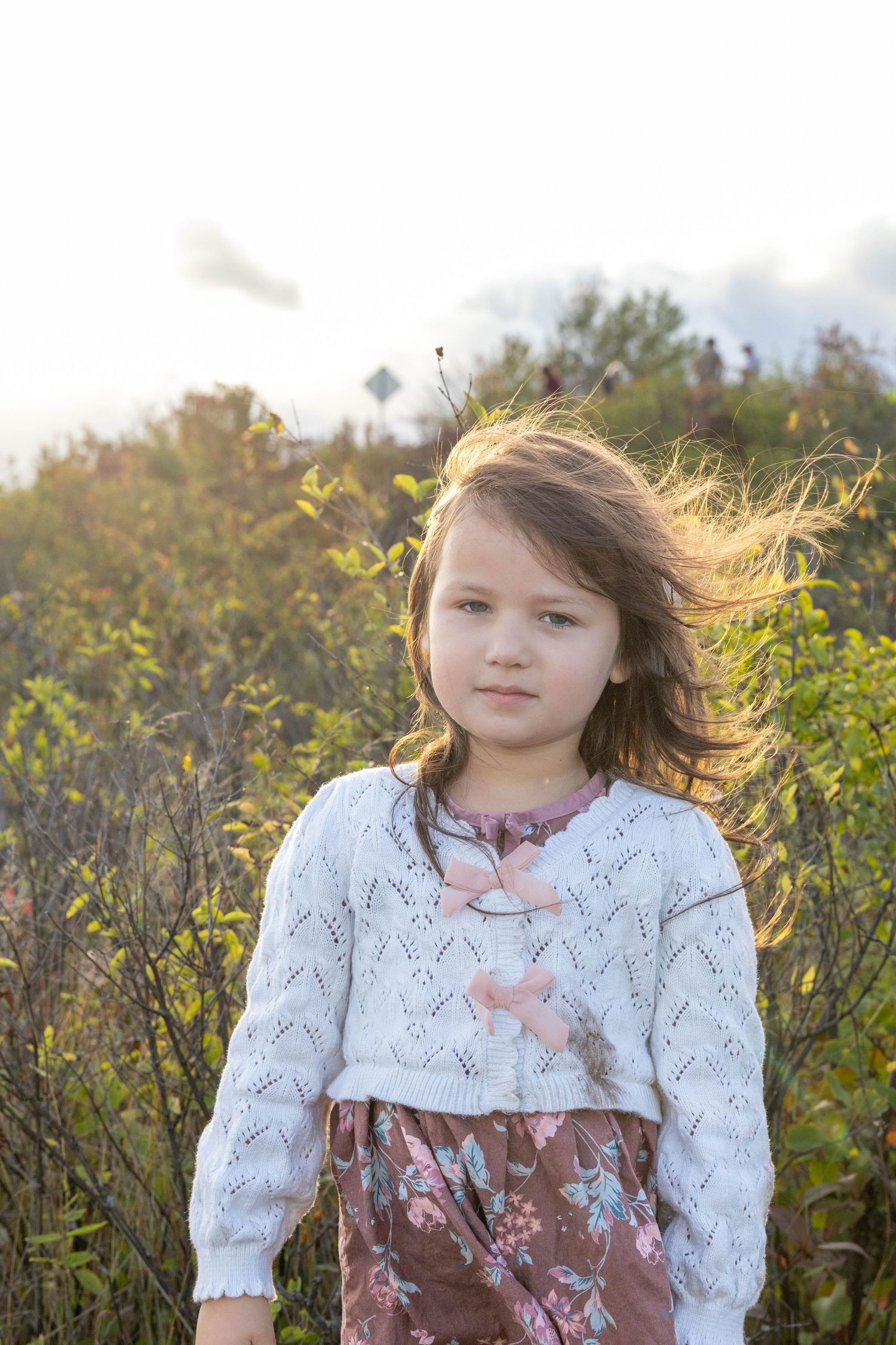 A child in a white lace top and pink bow poses in front of autumn foliage during a golden hour photoshoot.