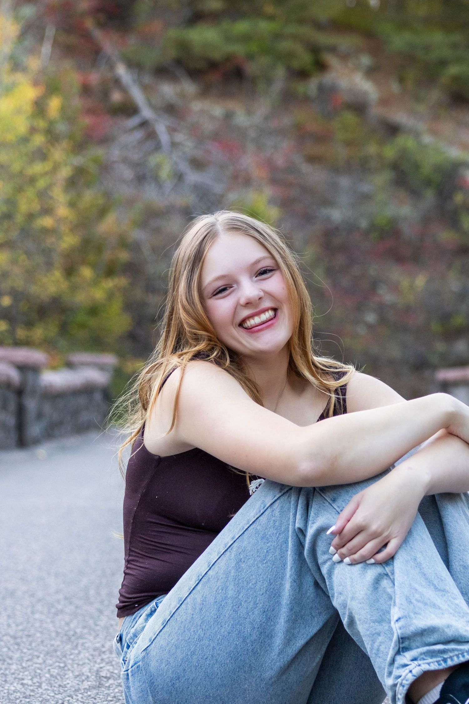 A joyful portrait taken outdoors showing someone sitting casually on pavement with autumn foliage in the background.